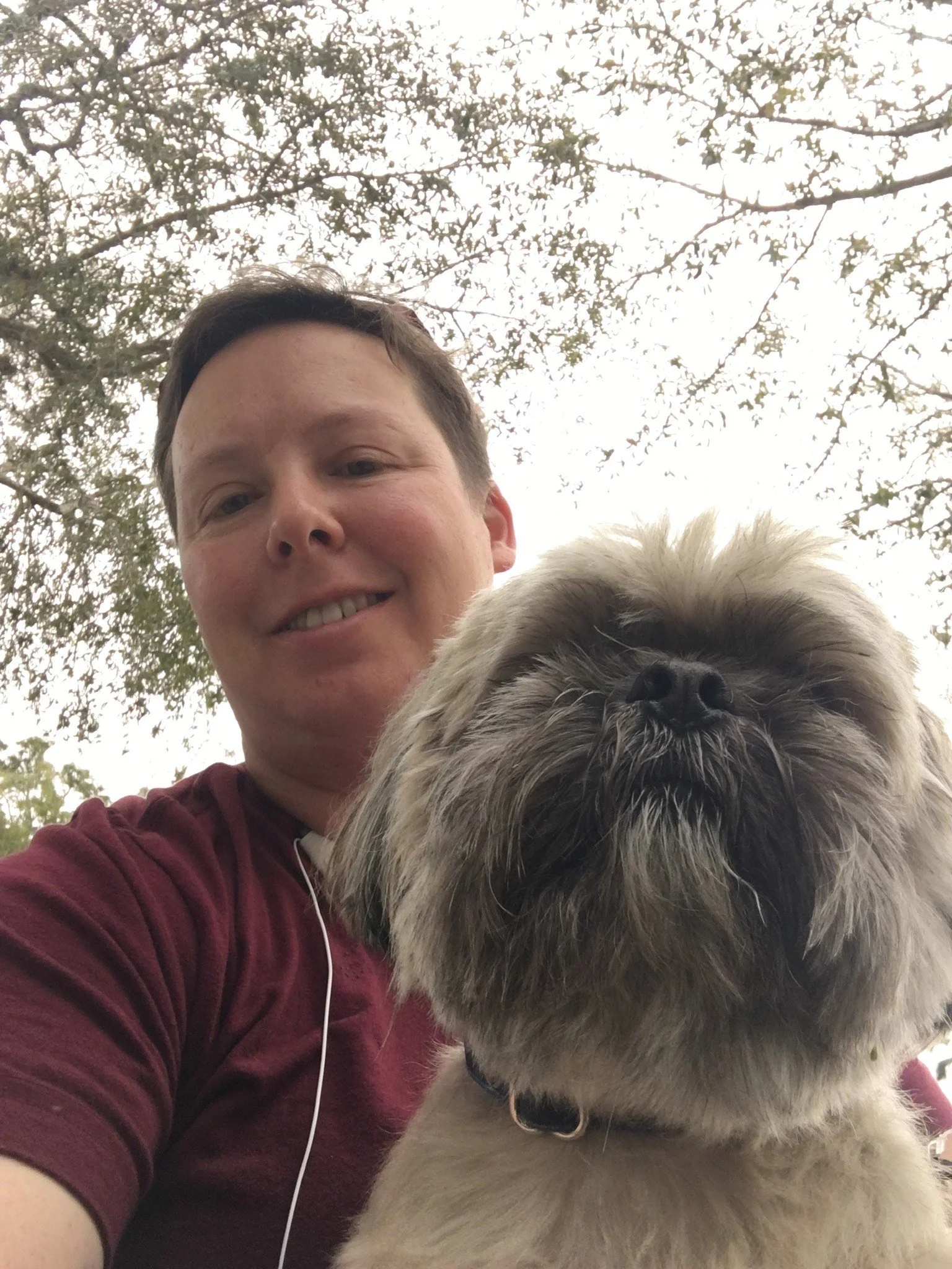 A person smiling and taking a selfie with a small, fluffy dog outdoors. The background features trees and a cloudy sky.