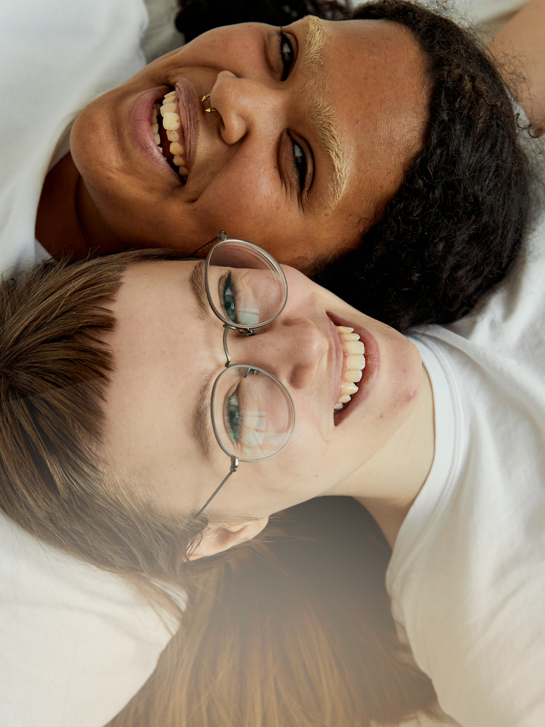 Two young women lie close together, smiling and looking upward in a relaxed, joyful moment. One wears glasses, while the other has braided hair and glasses, creating a warm and candid scene.