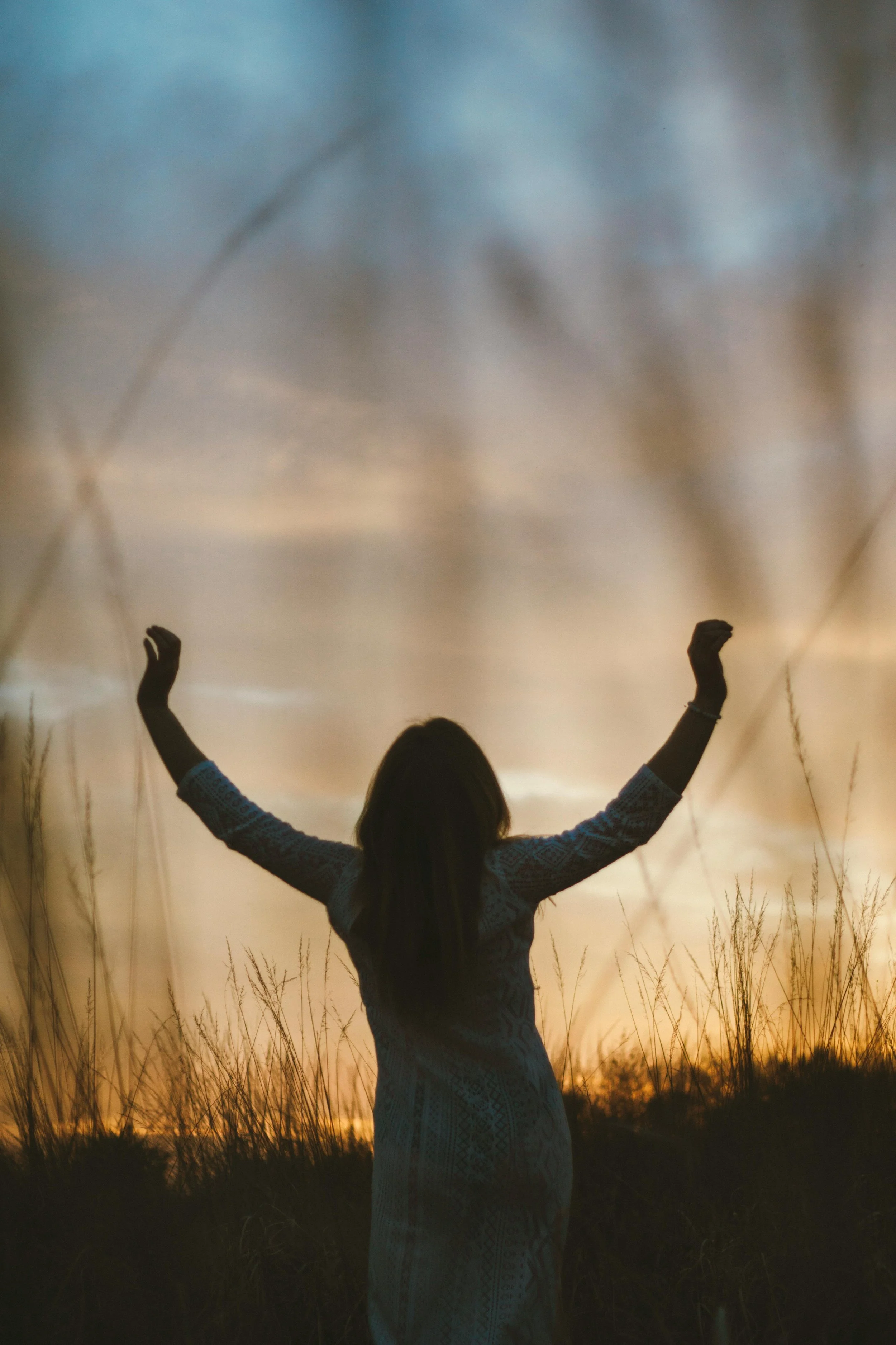 A girl standing in a field with tall grass at sunset, raising her arms with palms facing upward.