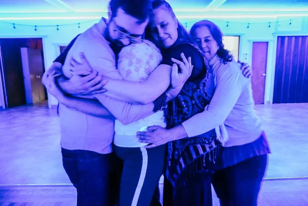 Four people embrace in a warm group hug at a lively gathering, with soft string lights glowing in the background, creating a cozy and heartfelt atmosphere.