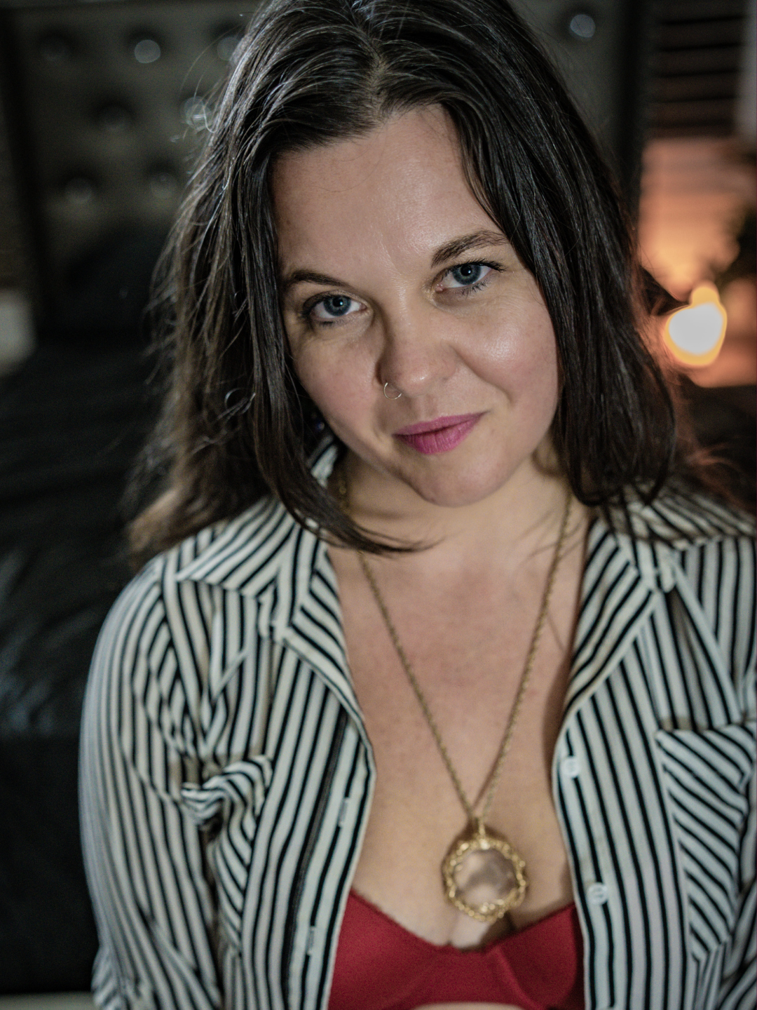 A quiet, cinematic portrait of a woman seated on a black leather couch, her layered outfit and pendant necklace adding texture against the soft, low-light ambiance.