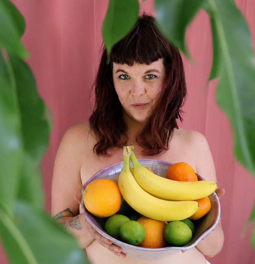 A woman with dark red hair holding a bowl of colorful fresh fruit, including bananas, oranges, and limes, with large green leaves partially framing the view and a pink background.