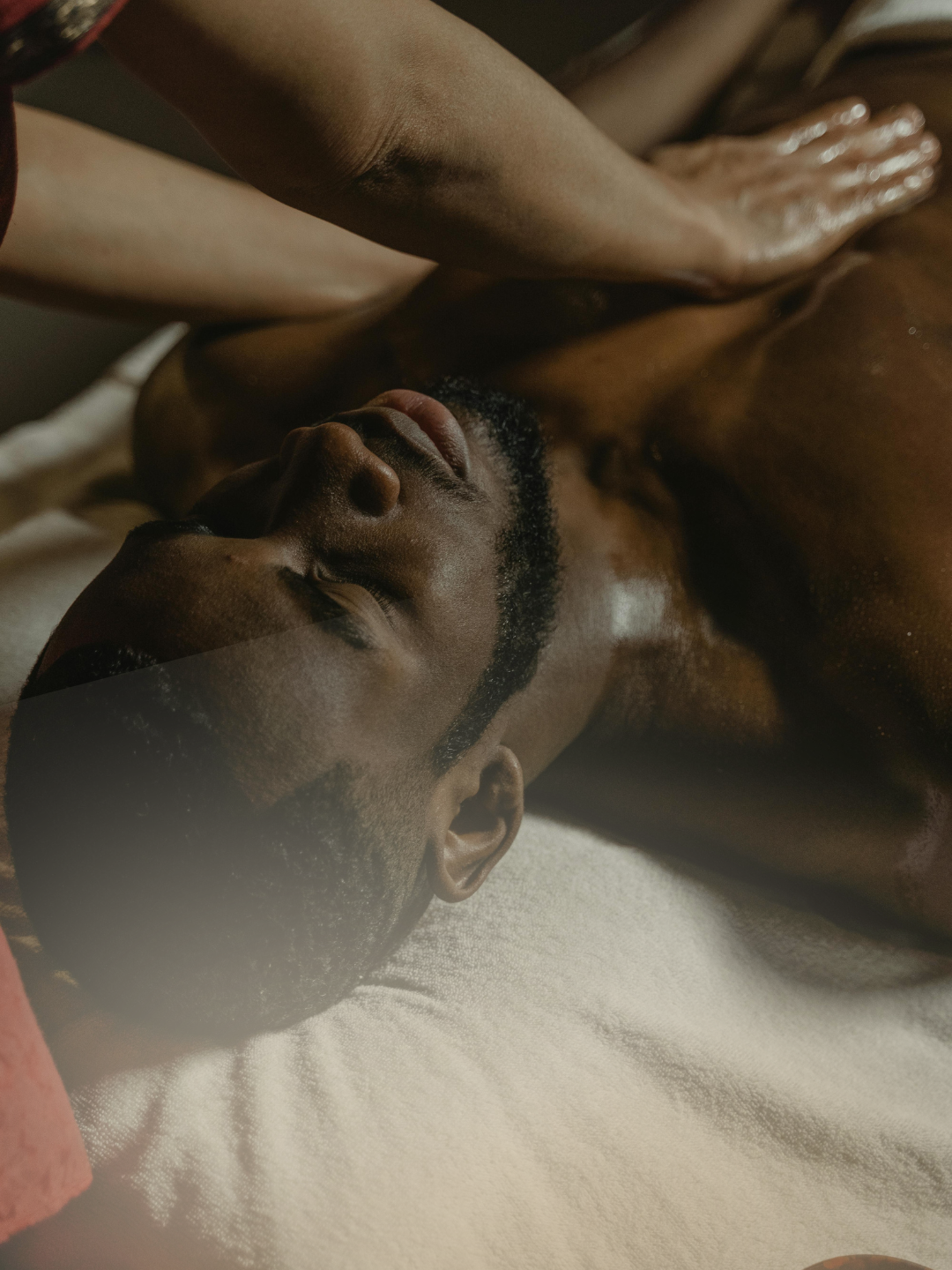 A dark-skinned man lies on a bed with eyes closed, one arm resting overhead and the other on his chest, captured in a calm, deeply relaxed moment against soft white sheets.