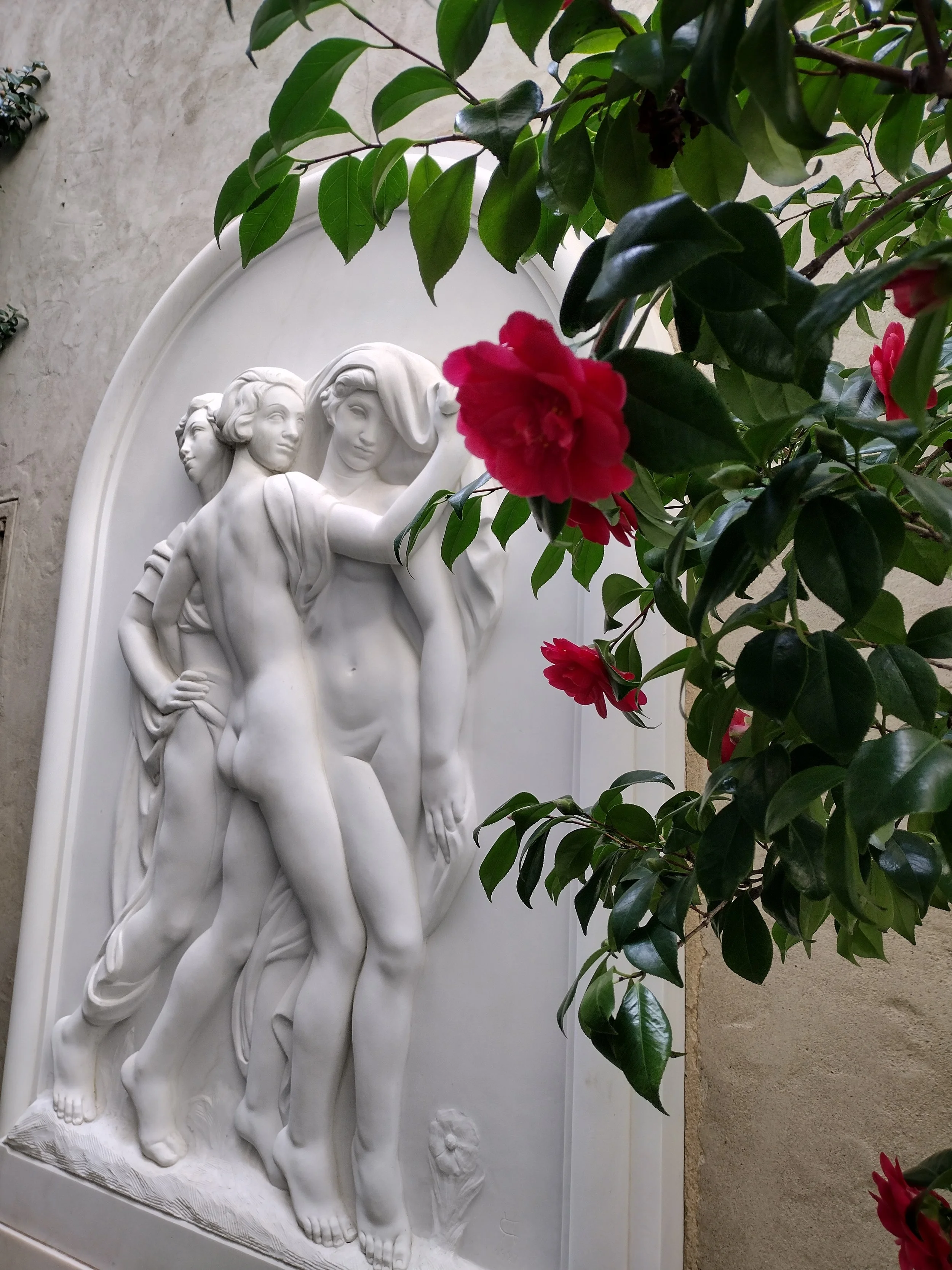 A white sculpted relief depicting three women standing side by side, partially framed by green leaves and soft pink flowers in the foreground, creating a layered and elegant composition.