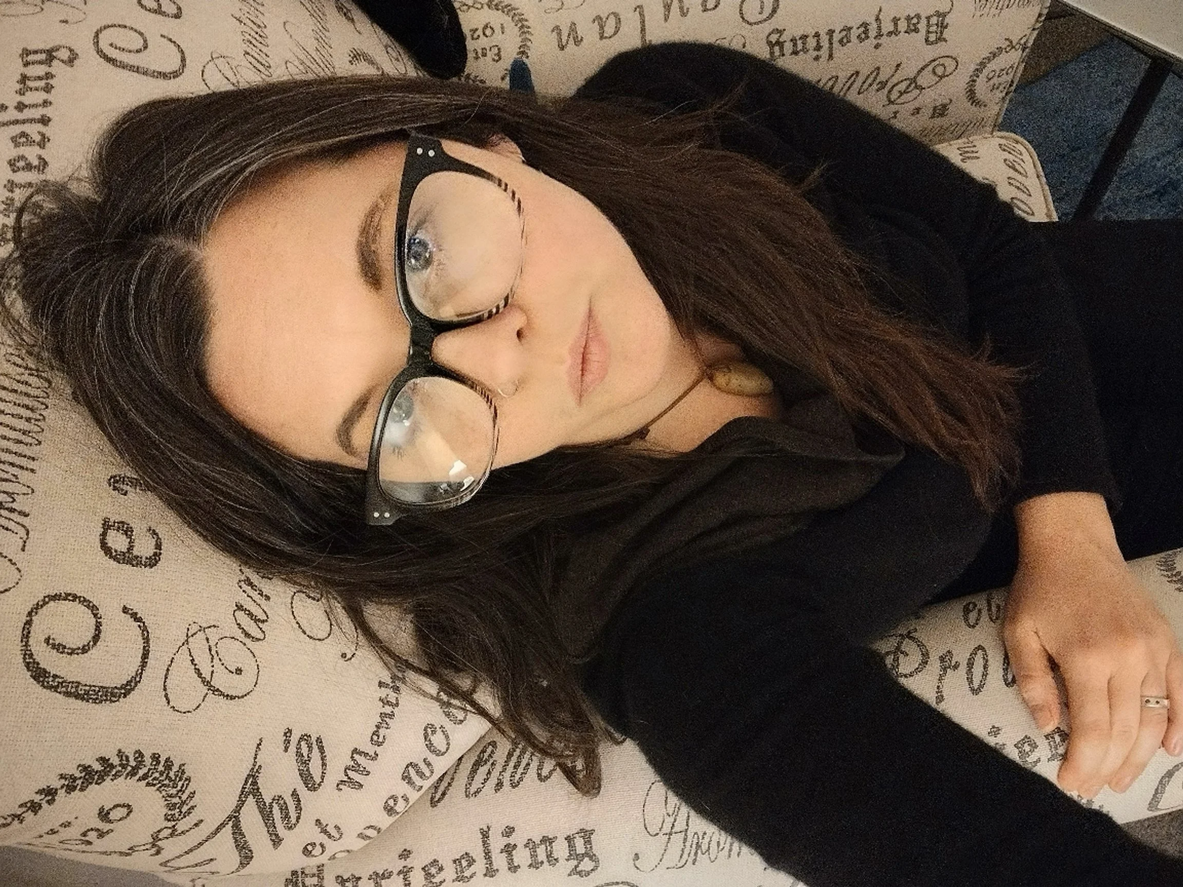 A woman with long brown hair, glasses, and a septum piercing lies on a cushion with printed words, smiling as she makes a peace sign with her hand.