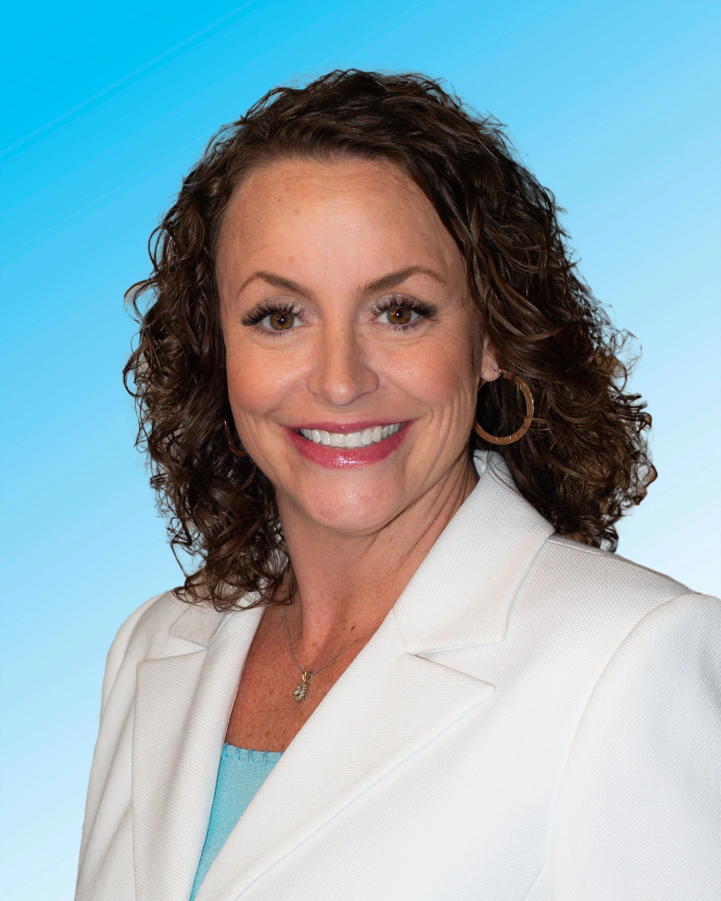 A woman with curly brown hair smiling, wearing a cream blazer and a necklace, against a gradient blue background.