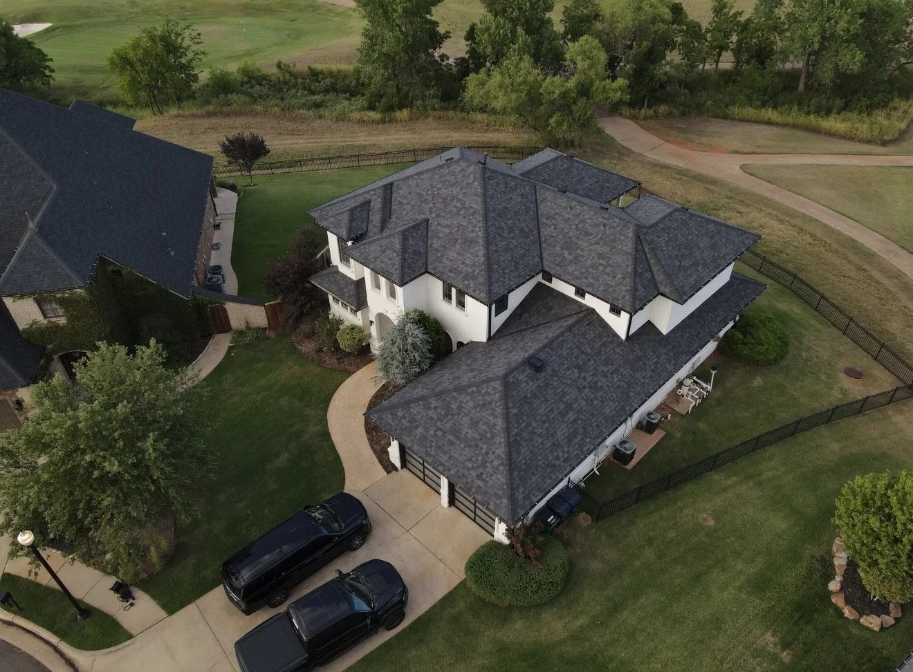 Aerial view of a large two-story suburban house with gray roof, white walls, driveway with three parked cars, surrounded by green lawn, trees, and a fenced backyard.