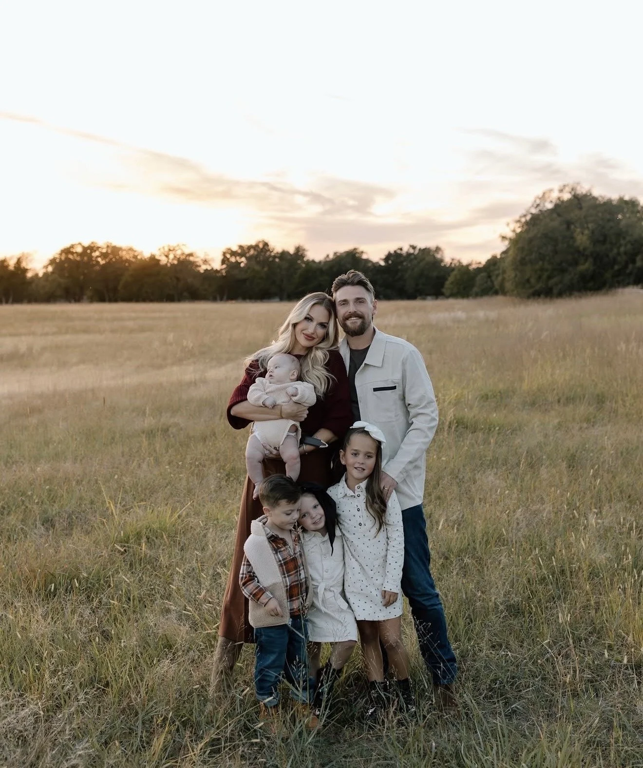 Family of six posing in a field at sunset, including two parents, three children, and a baby, with trees and a colorful sky in the background.