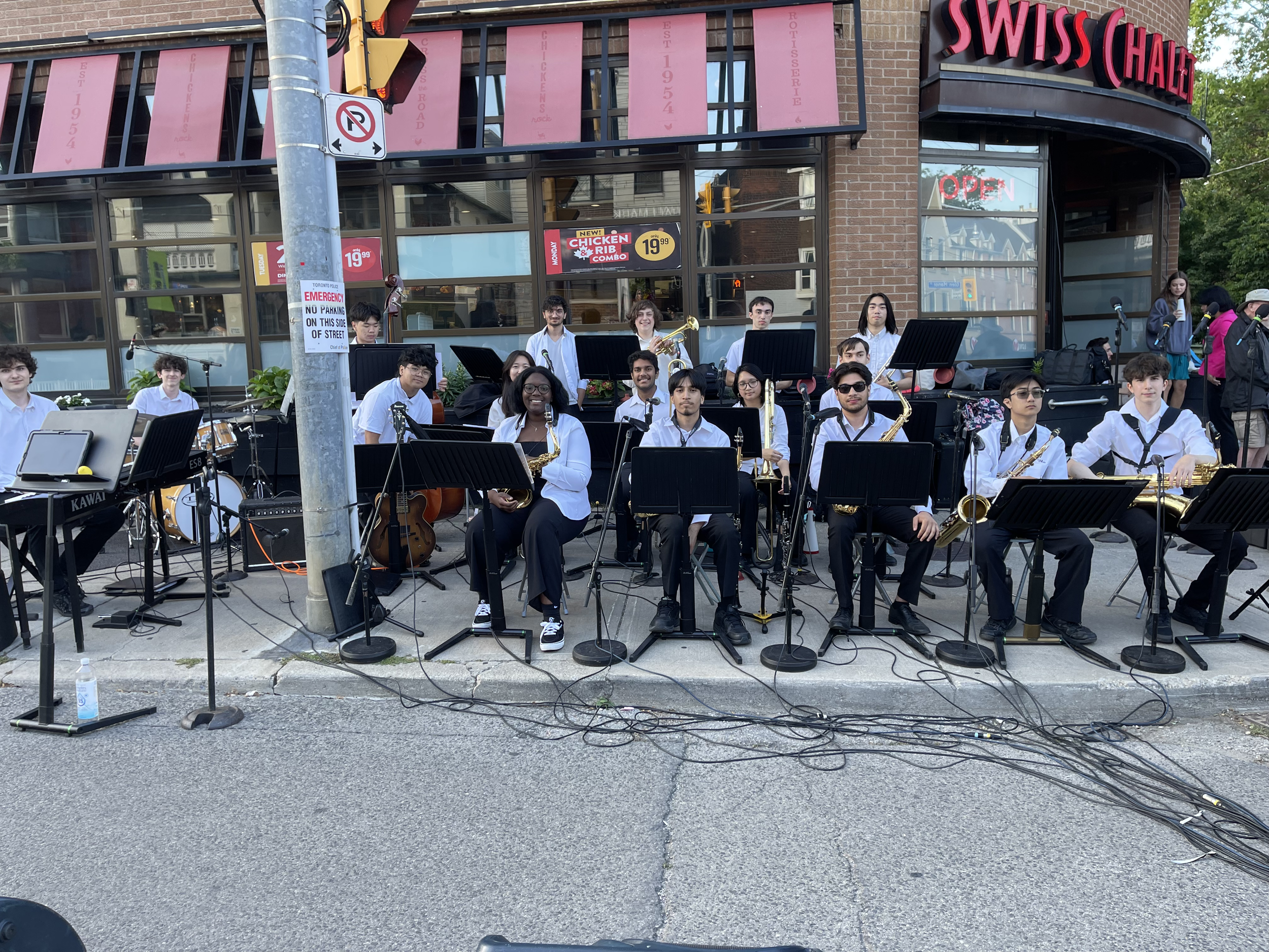 A group of musicians with instruments, sitting and standing on a city sidewalk outside a restaurant called Swiss Chalet, with some onlookers in the background.