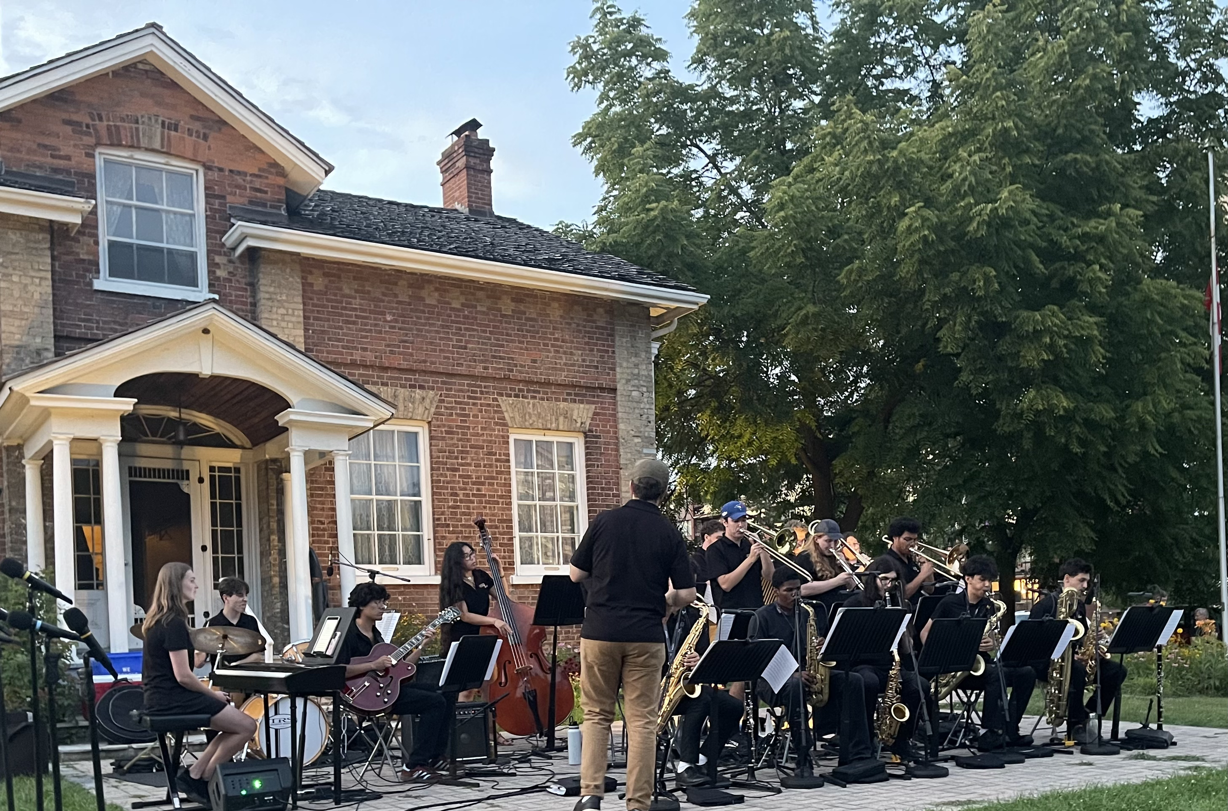 A large outdoor musical performance with a band of musicians playing instruments like saxophones, guitars, drums, and a keyboard, in front of a brick house with large windows, trees, and a flagpole.