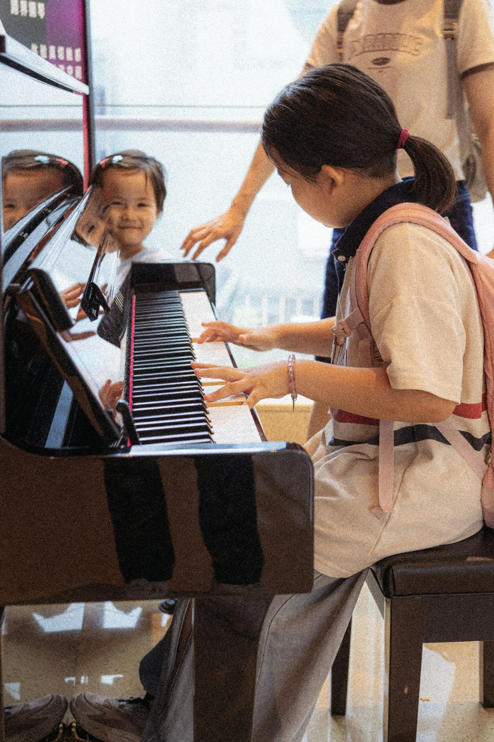 A young girl with a ponytail playing piano, smiling at her reflection in the piano keys, while an adult assists her.