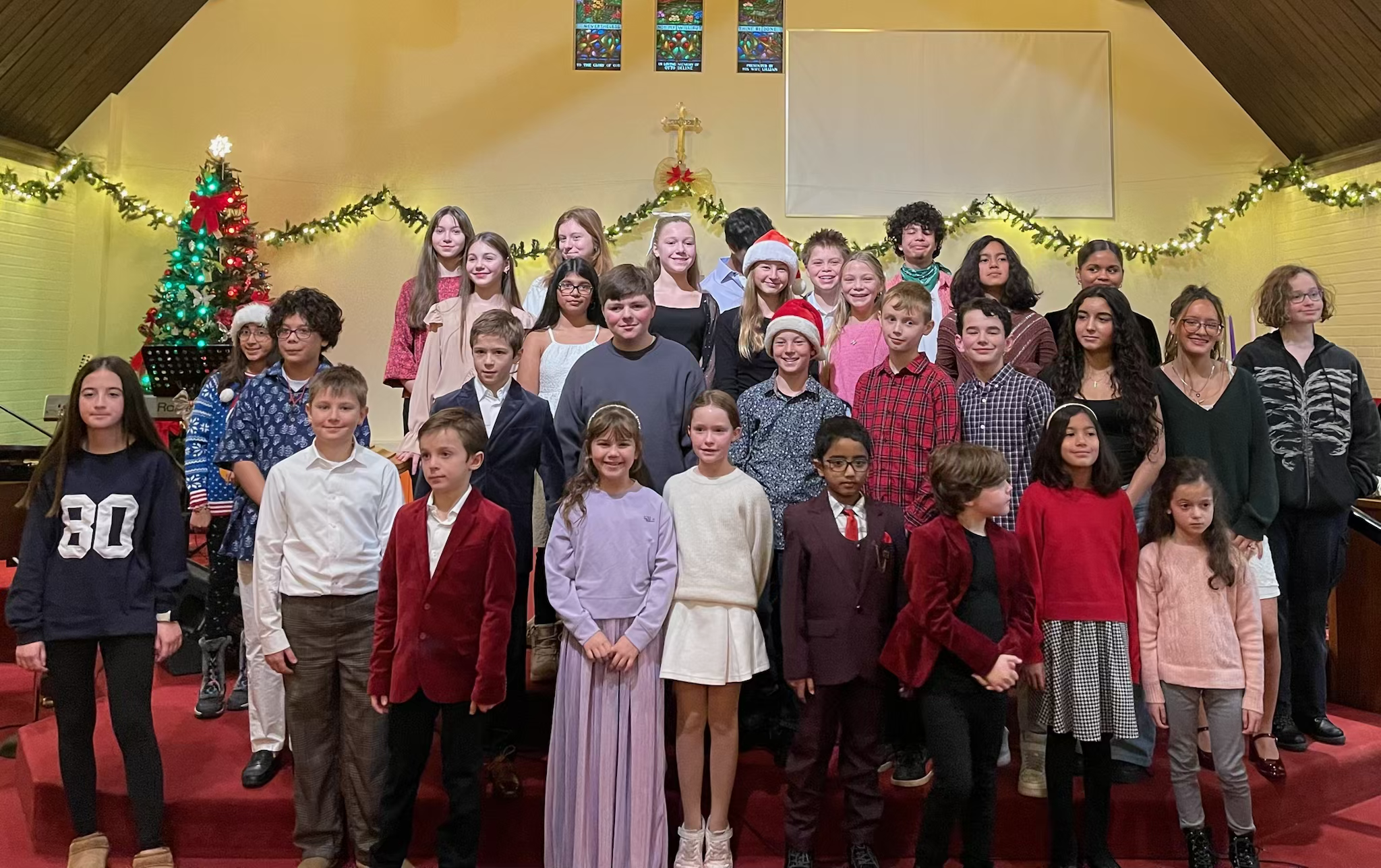 A group of children and teenagers standing on a stage inside a church, celebrating Christmas. Some are wearing Santa hats, and there is a decorated Christmas tree with lights and ornaments to the left. The background has a yellow wall with green garl