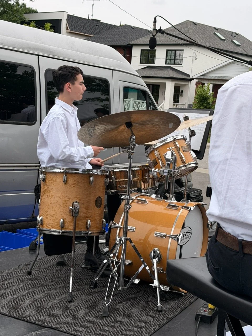 Young man playing drums outdoors, with a house and van in the background, and a boom microphone overhead.