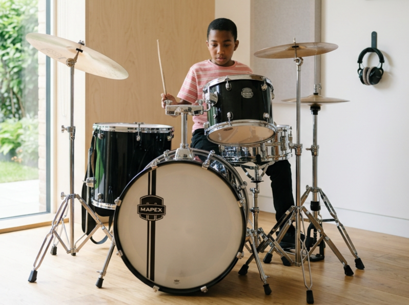 A young boy sitting behind a black drum set in a room with wooden floor, natural light, and headphones hanging on the wall.