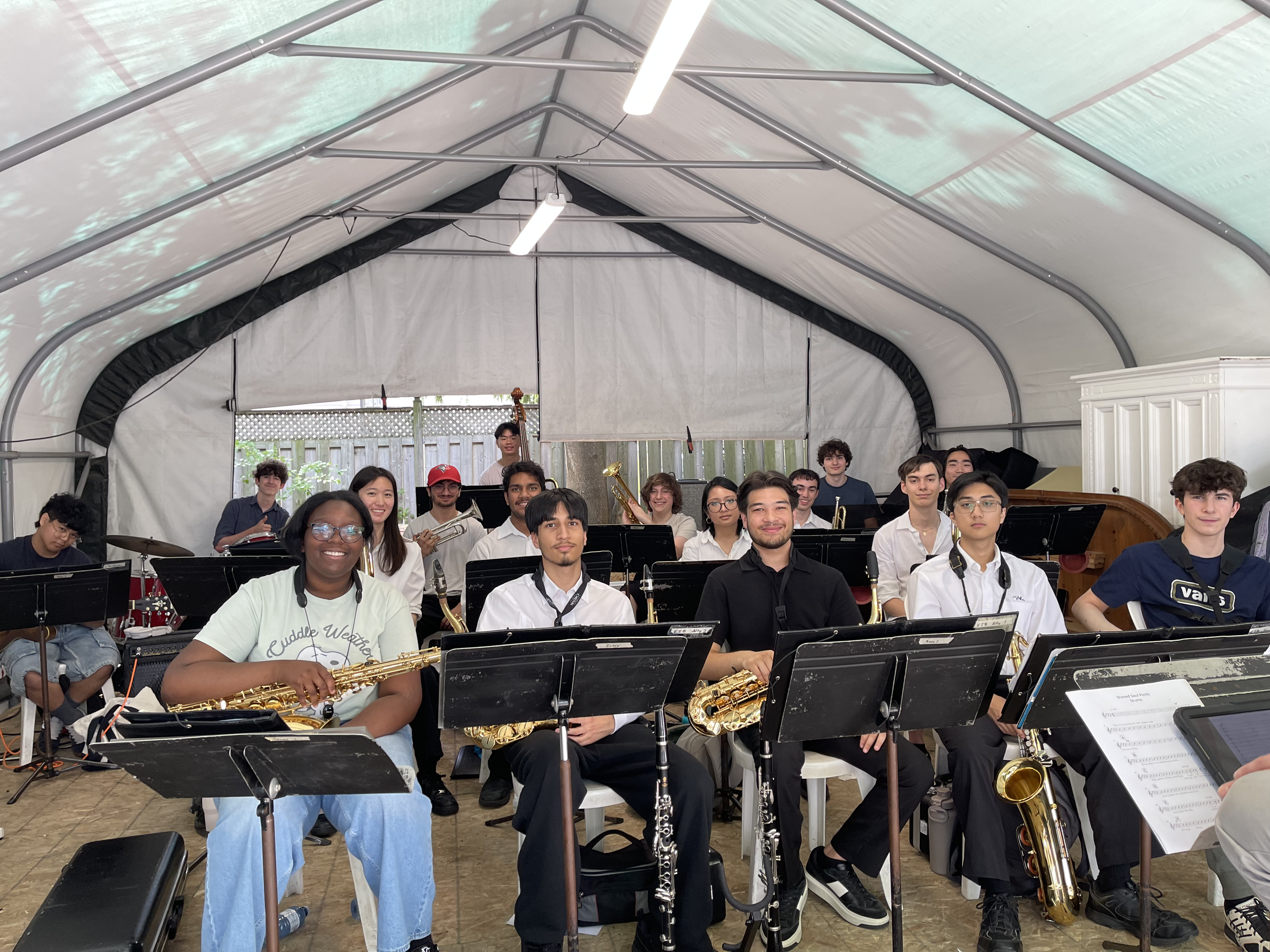 A group of diverse young people in a covered outdoor space, some holding musical instruments like saxophones, with music stands and sheet music in front.