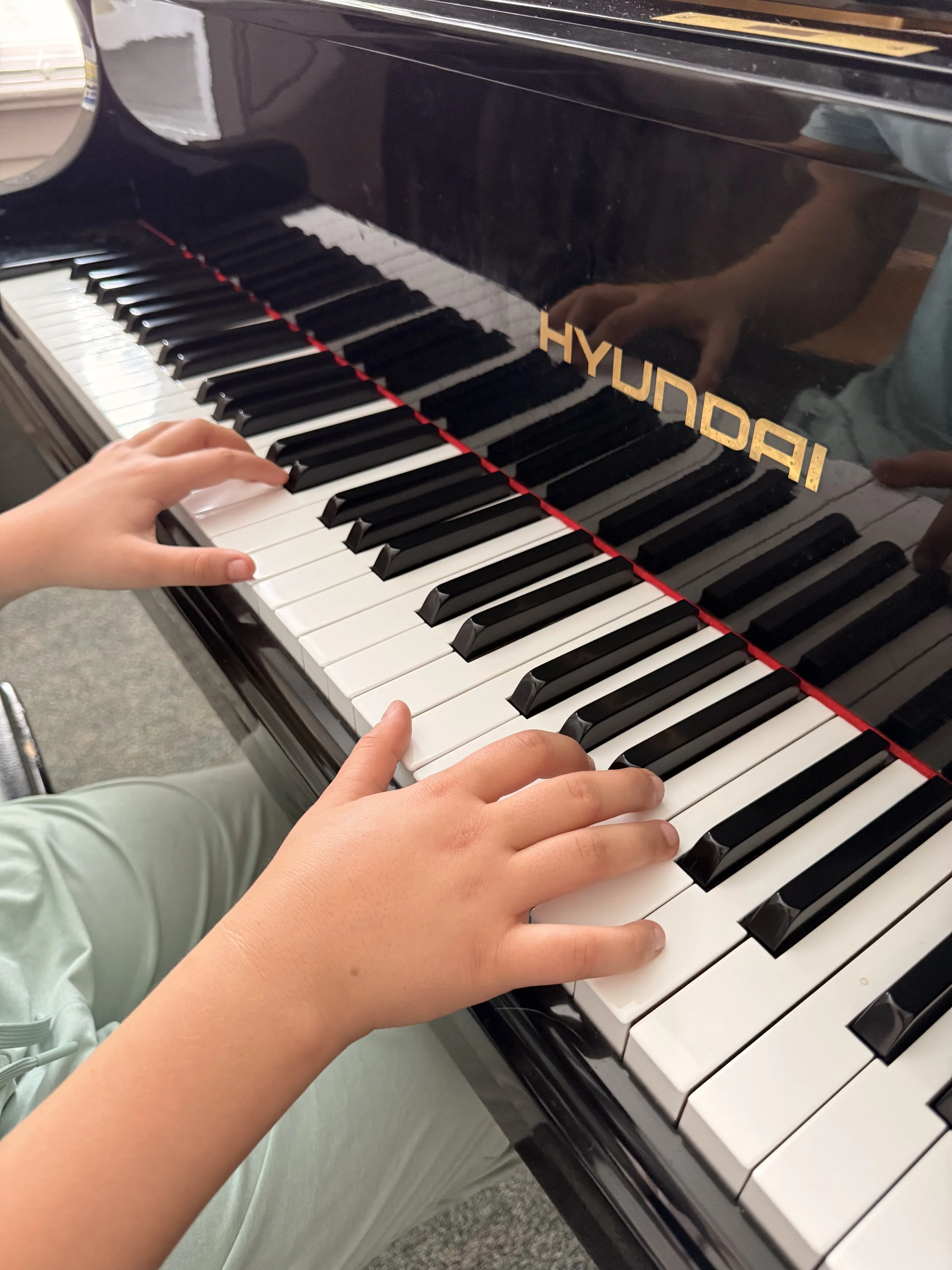 A child's hands playing the keys of a black grand piano with the brand name 'HYUNDAI' visible on the side.