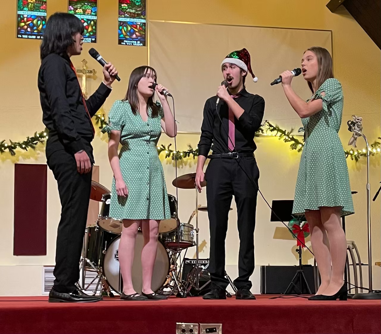 Four people singing on a stage decorated with Christmas garland. One man wears a Santa hat. The three women are in green polka dot dresses. There is a drum set behind them.