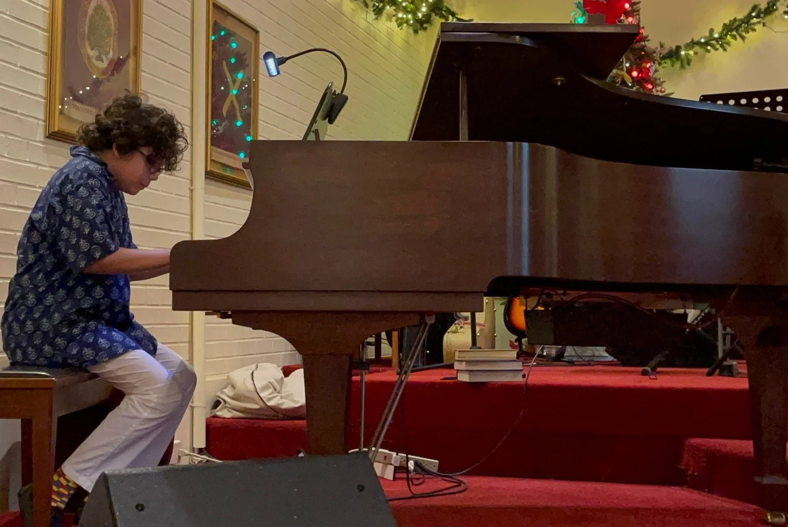 A person with curly hair wearing a blue patterned shirt and white pants, sitting on a chair and playing a grand piano in a decorated room with holiday lights and pictures on the wall.