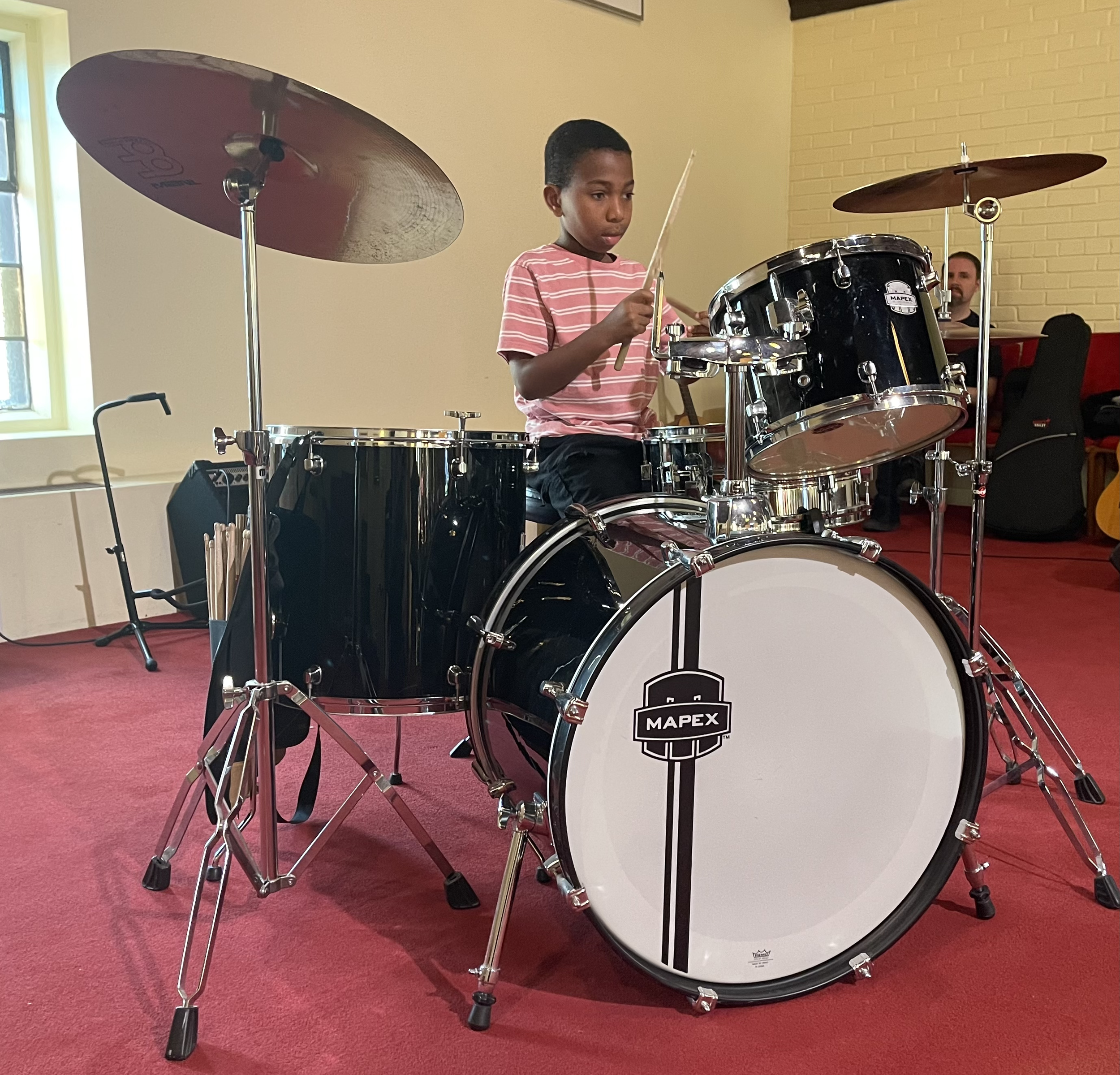 A young boy playing a black drum set with silver hardware, with a serious expression, in a room with yellow brick walls and red carpet. There are two drums, two cymbals, and a music stand nearby.