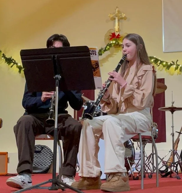 Two people playing clarinets on a decorated stage with Christmas decor including a cross, a poinsettia, and garlands.