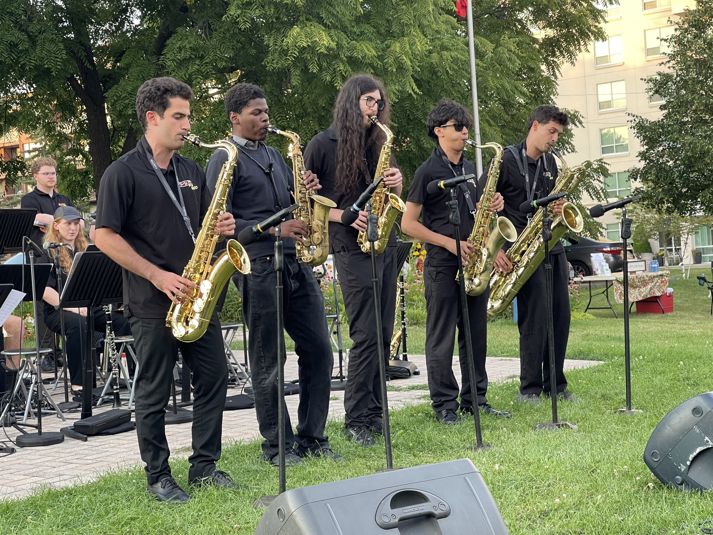 Five musicians, dressed in black, playing saxophones at an outdoor event, with trees, a building, and a woman in a cap in the background.