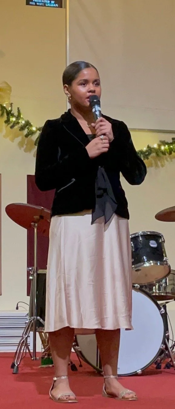 A woman with dark hair pulled back, wearing a black velvet jacket, beige satin skirt, and open-toed heels, standing on a stage with a microphone in her hand, with musical instruments and holiday decorations in the background.