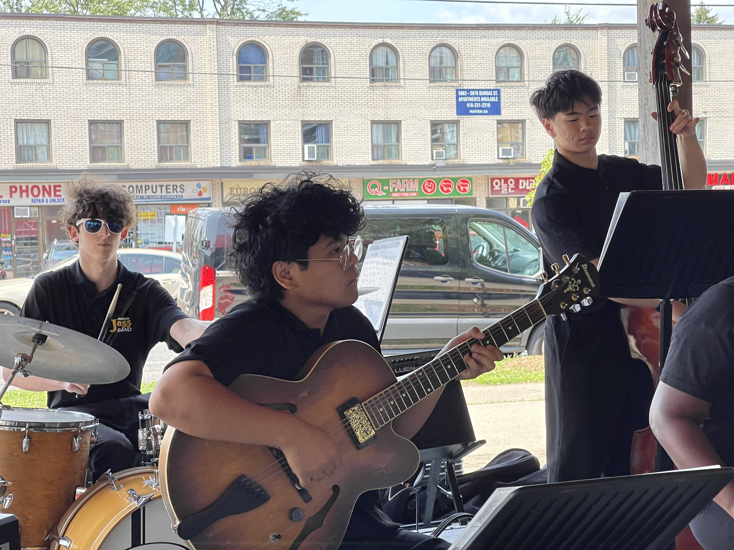 A group of musicians playing instruments outdoors. One person is playing drums, another is playing the guitar, and a third person is playing the bass. The background shows a brick building with stores and parked cars.