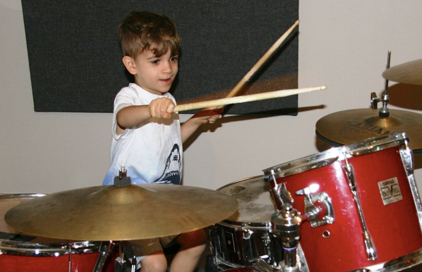 A young boy playing a red drum set, holding drumsticks in both hands, with a focused expression.