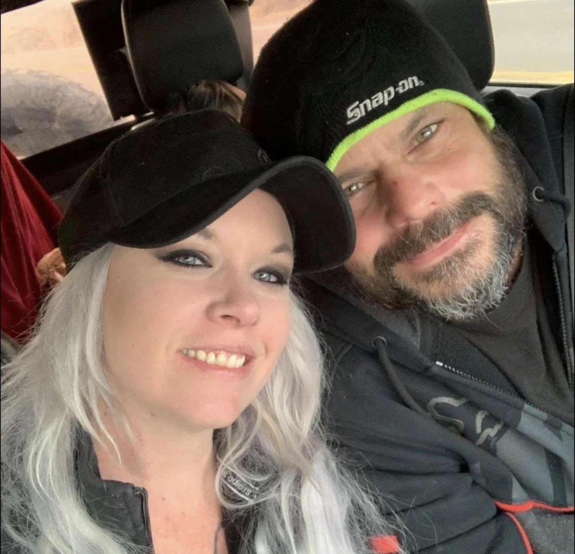 A smiling woman with long gray hair and a black cap sitting next to a smiling man with a beard wearing a black beanie and hoodie inside a vehicle.