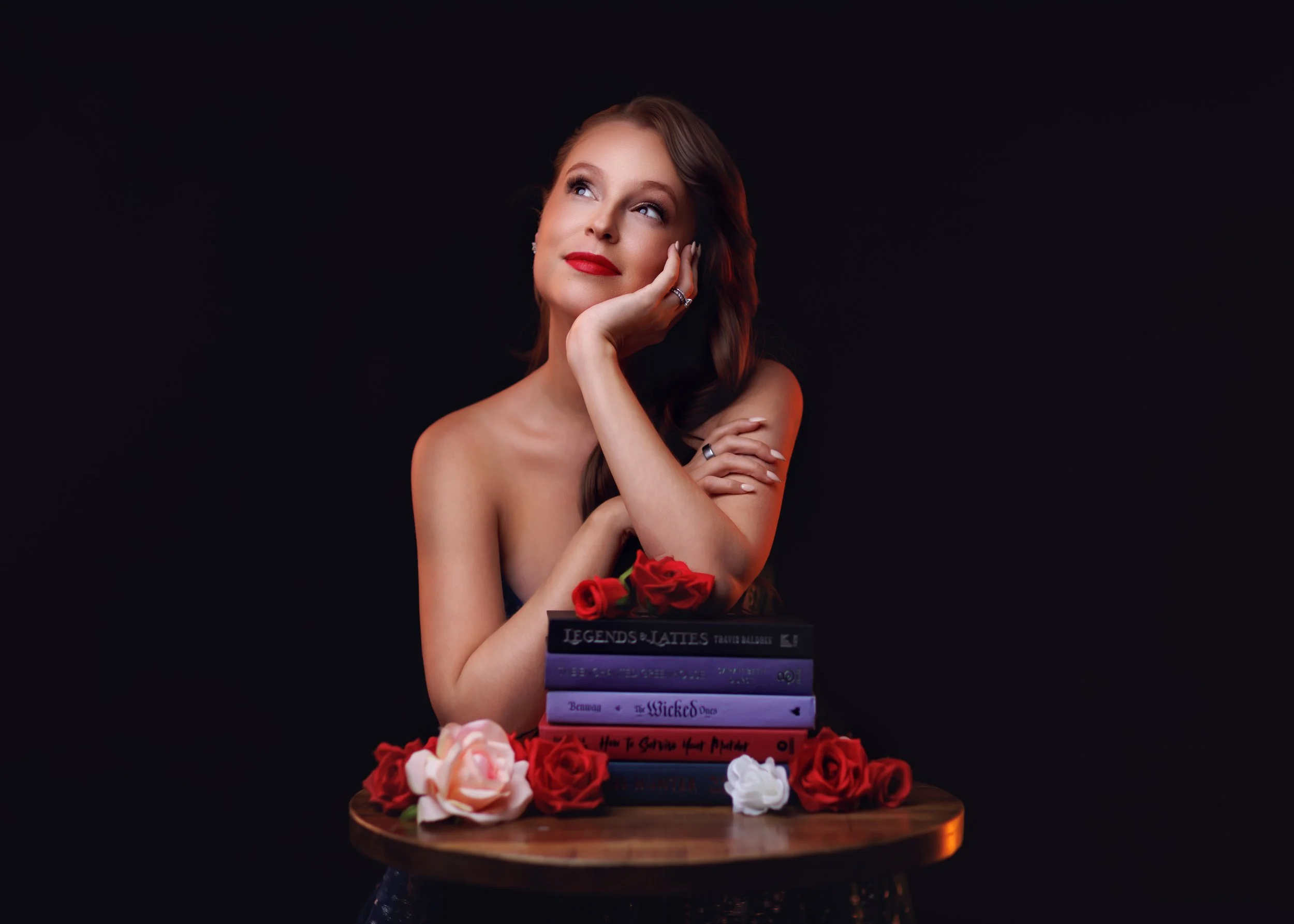 A woman with long brown hair and red lipstick resting her face on her hand, sitting behind a stack of books with red and white roses on top, against a dark background.