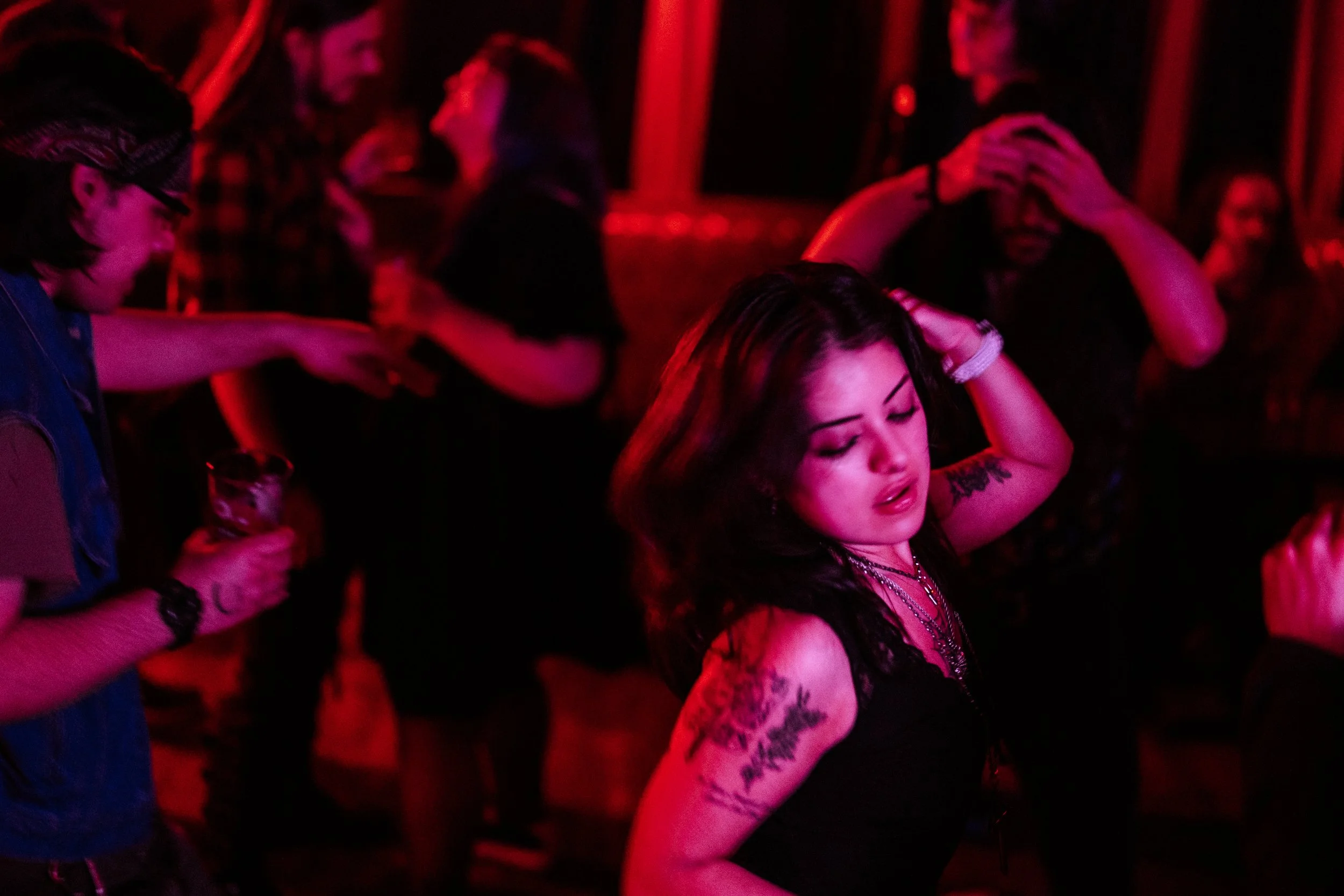 A group of young women dancing in a dimly lit nightclub with red lighting, some wearing glasses and tattoos, enjoying the music.