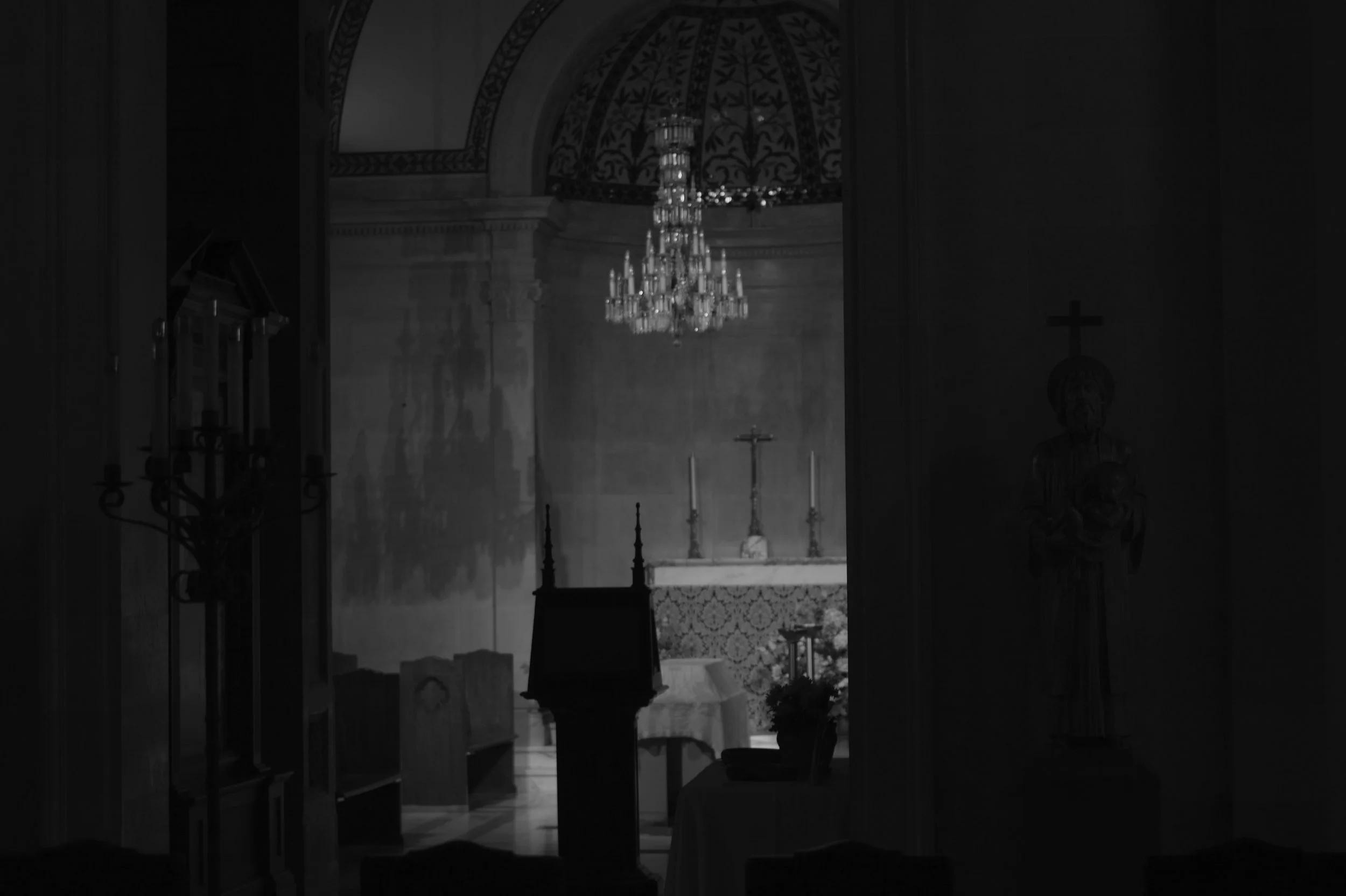 Dark interior of a church with a chandelier hanging from the ceiling, altars, and religious statues, with a cross in the background.