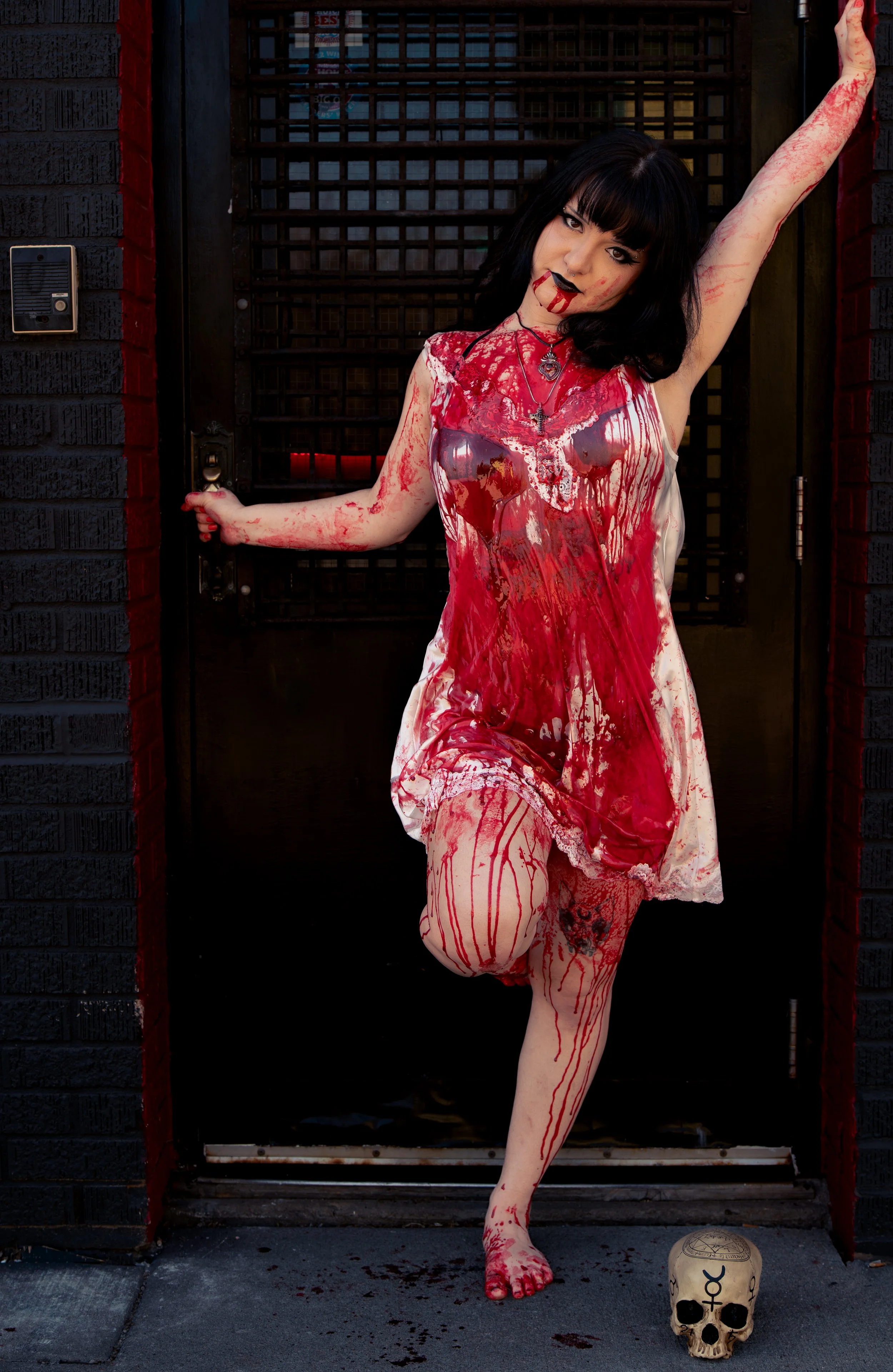 A woman with black hair and tattoos, dressed in a blood-stained dress, standing against a dark door with one arm raised and the other holding the door handle. She has fake blood on her face and hands and is positioned next to a skull with occult symbols on it, on a dark sidewalk.