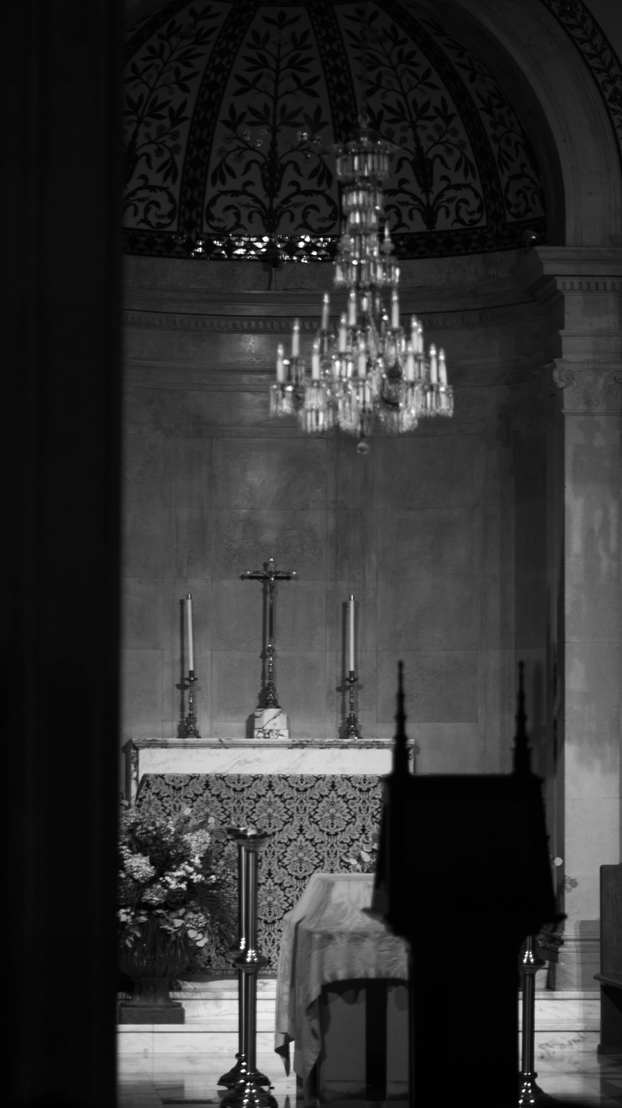 A black and white photo of a church altar with a cross, candles, a table covered with a cloth, and floral arrangements. There is a chandelier hanging from the ceiling.