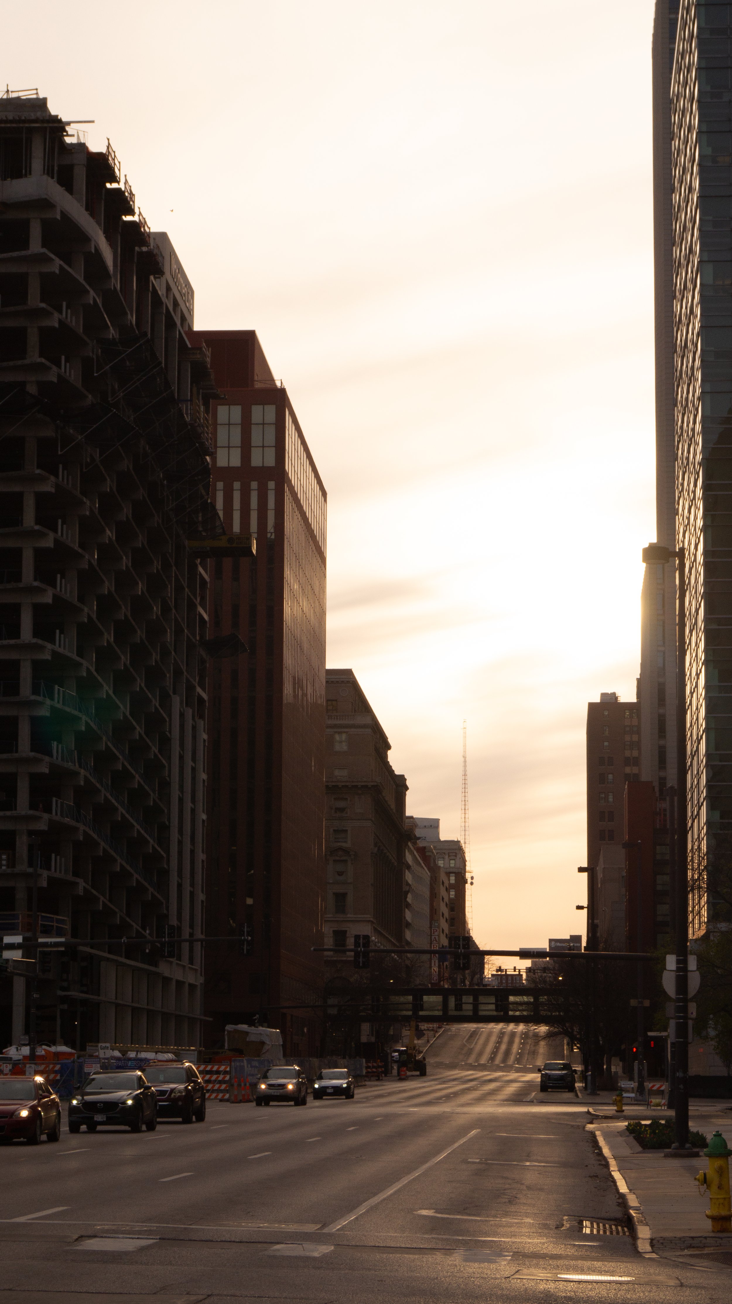 A city street during sunset with tall buildings on either side, some under construction, and cars on the road.