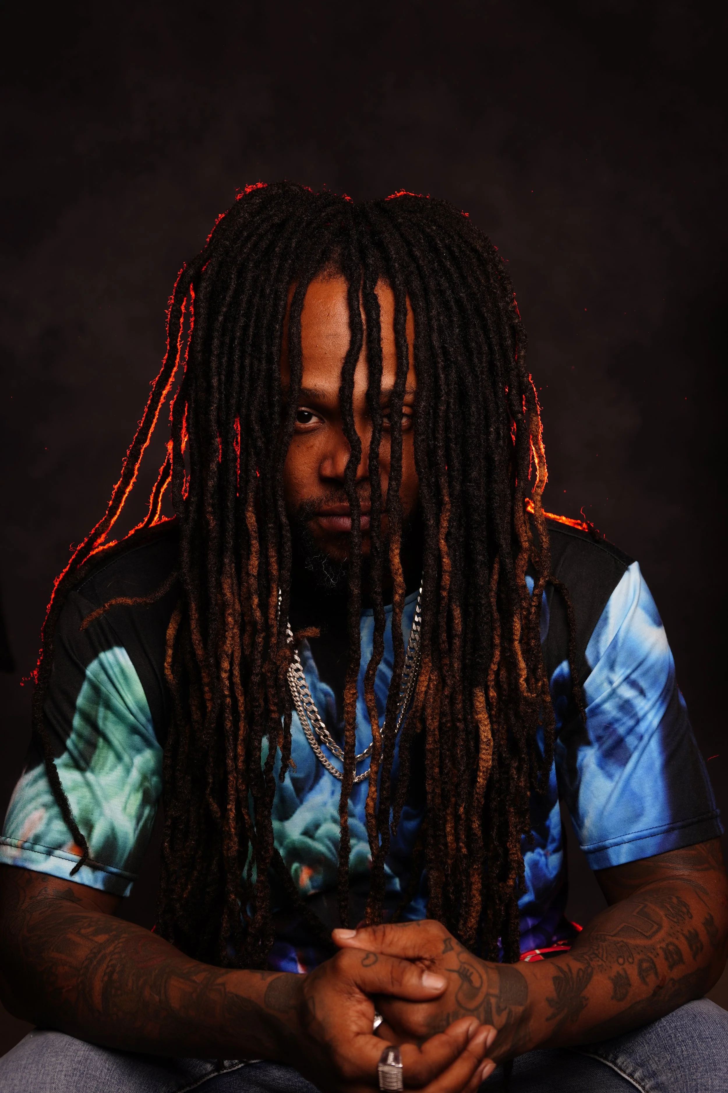 A person with dreadlocks and tattoos sits with arms crossed, wearing a colorful tie-dye shirt and multiple chains, against a dark background with red lighting accents.