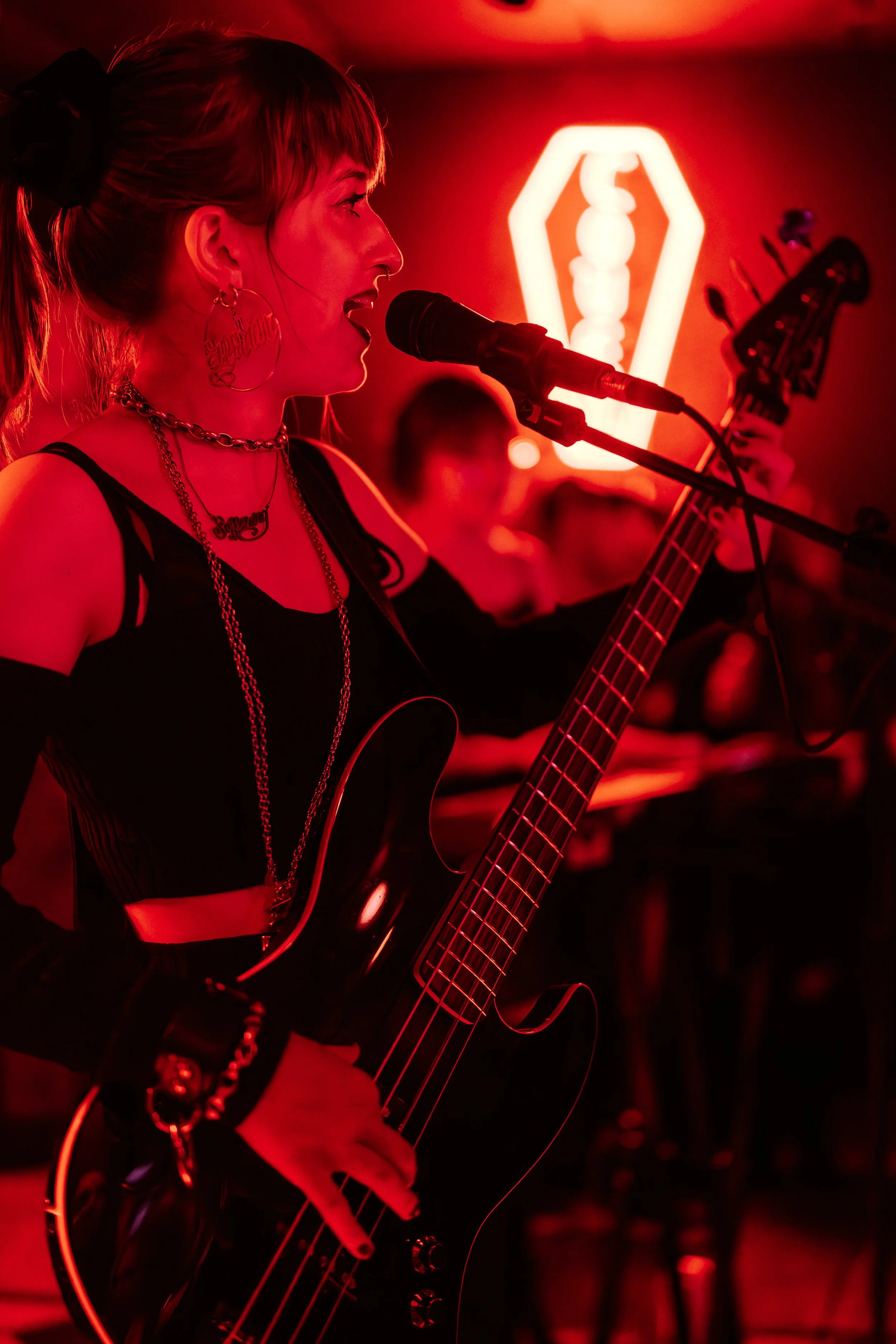 A woman with red hair and jewelry is singing into a microphone while playing an electric guitar, illuminated by red lighting and a neon sign in the background.