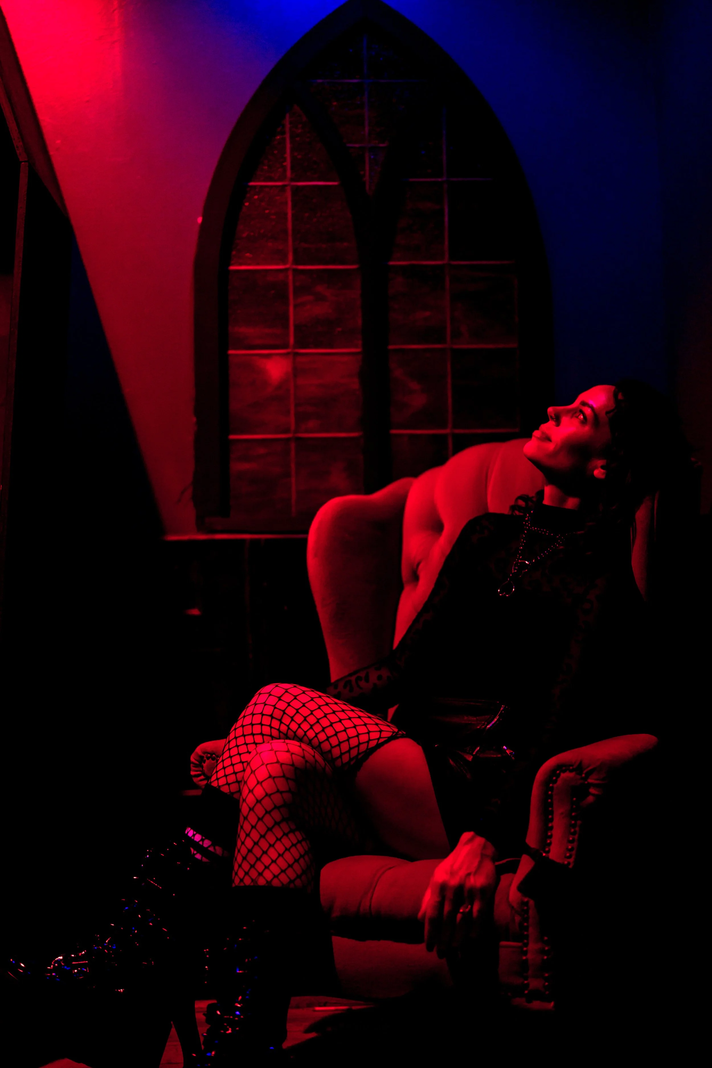 A woman with dark hair wearing fishnet stockings and a black outfit seated on a vintage armchair inside a room with a Gothic-style arched window, illuminated by red and blue lighting.