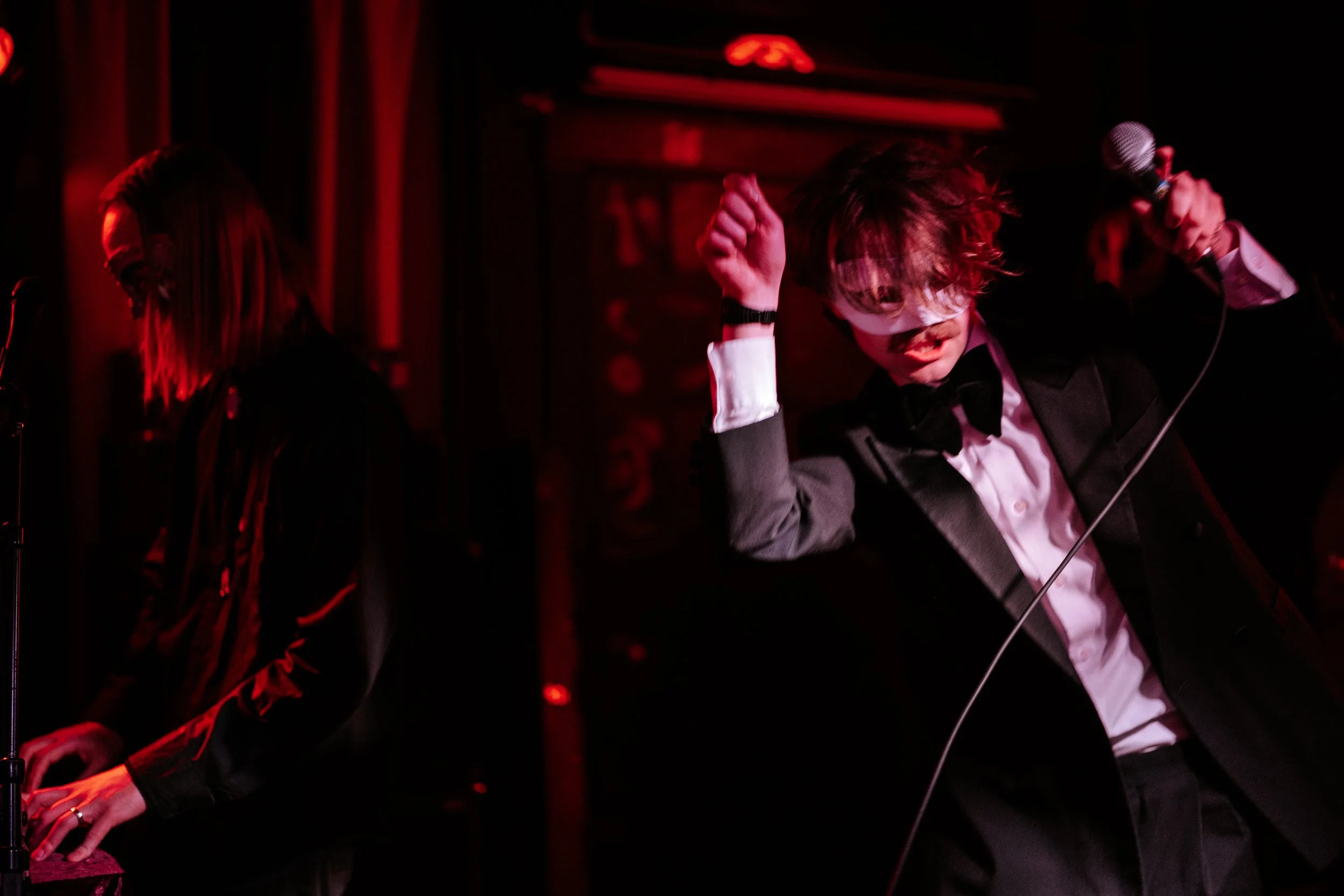 A man in a tuxedo wearing a blindfold, holding a microphone, and dancing on stage with red lighting in the background.