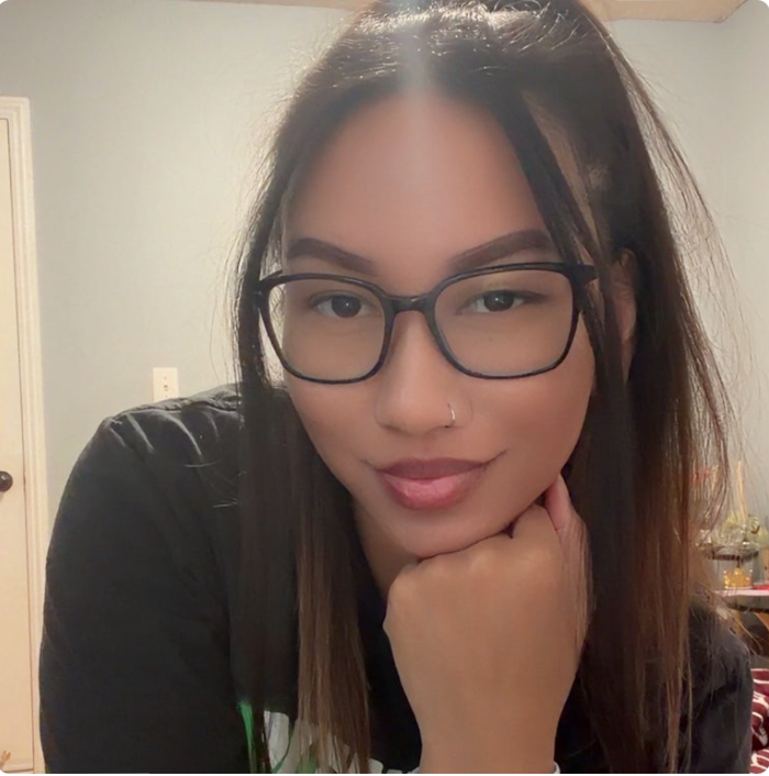 A young woman with long brown hair, wearing glasses with dark frames, a nose ring, and a black shirt, resting her chin on her hand, looking at the camera with a slight smile in an indoor setting.