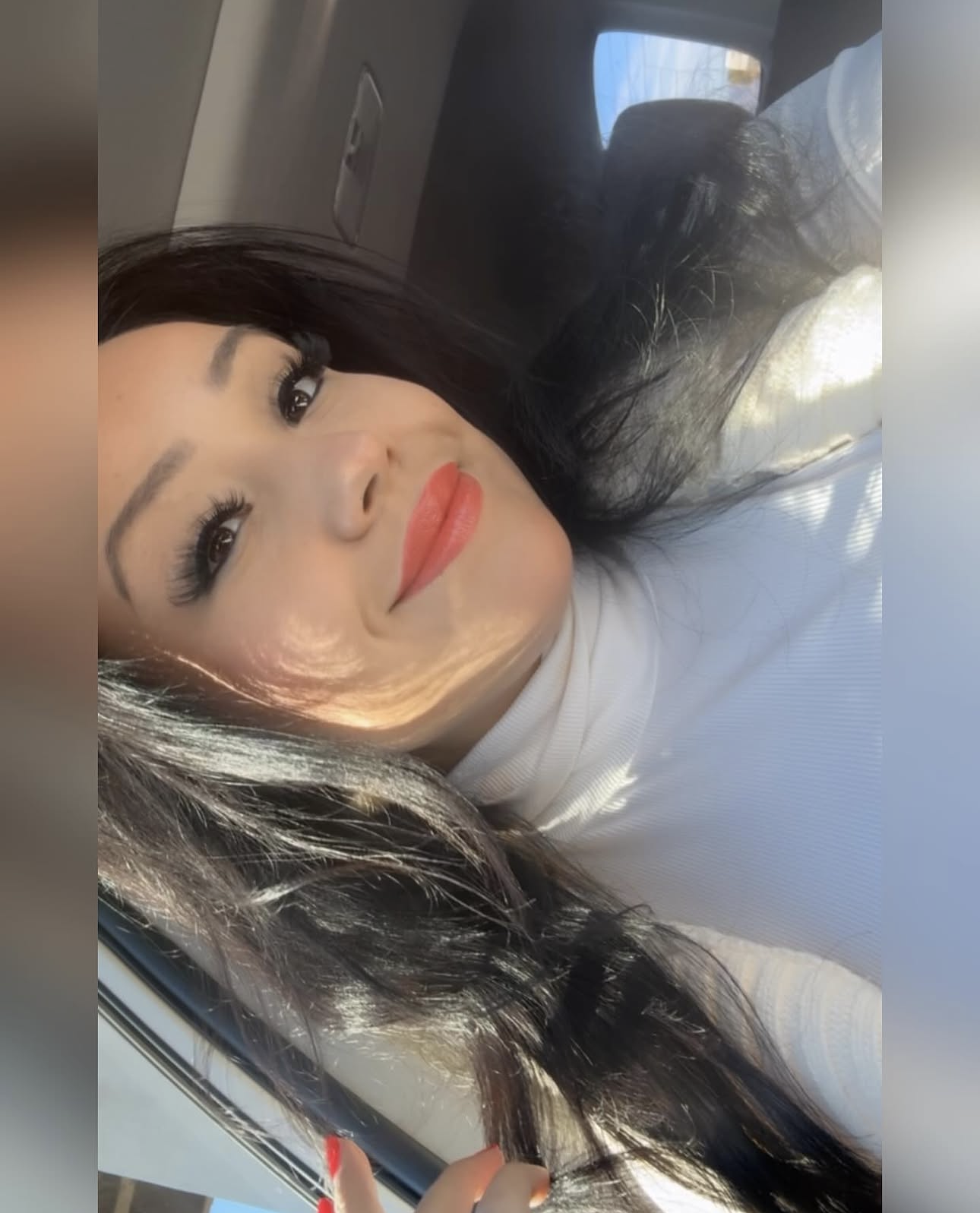 A woman with long black hair, makeup including eyeliner and red lipstick, taking a selfie inside a vehicle with sunlight streaming in.