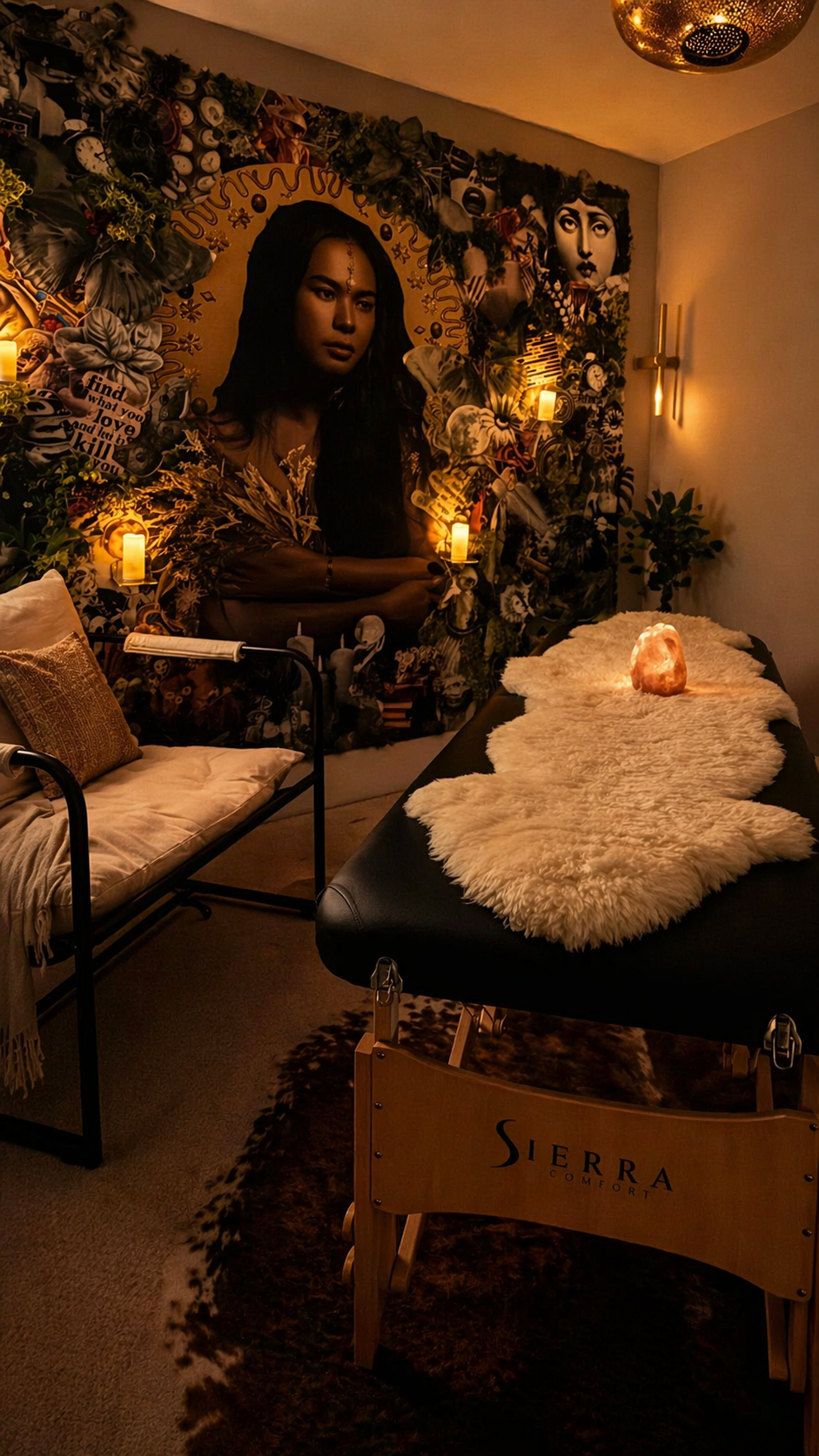 A cozy room decorated with a large, artistic mural of a woman with dark hair, surrounded by foliage, candles, and artwork. There is a massage table with a salt lamp, a plush sheepskin, and a sign that says "Sierra Comfort" on it. The scene is warmly lit with a calming ambiance.