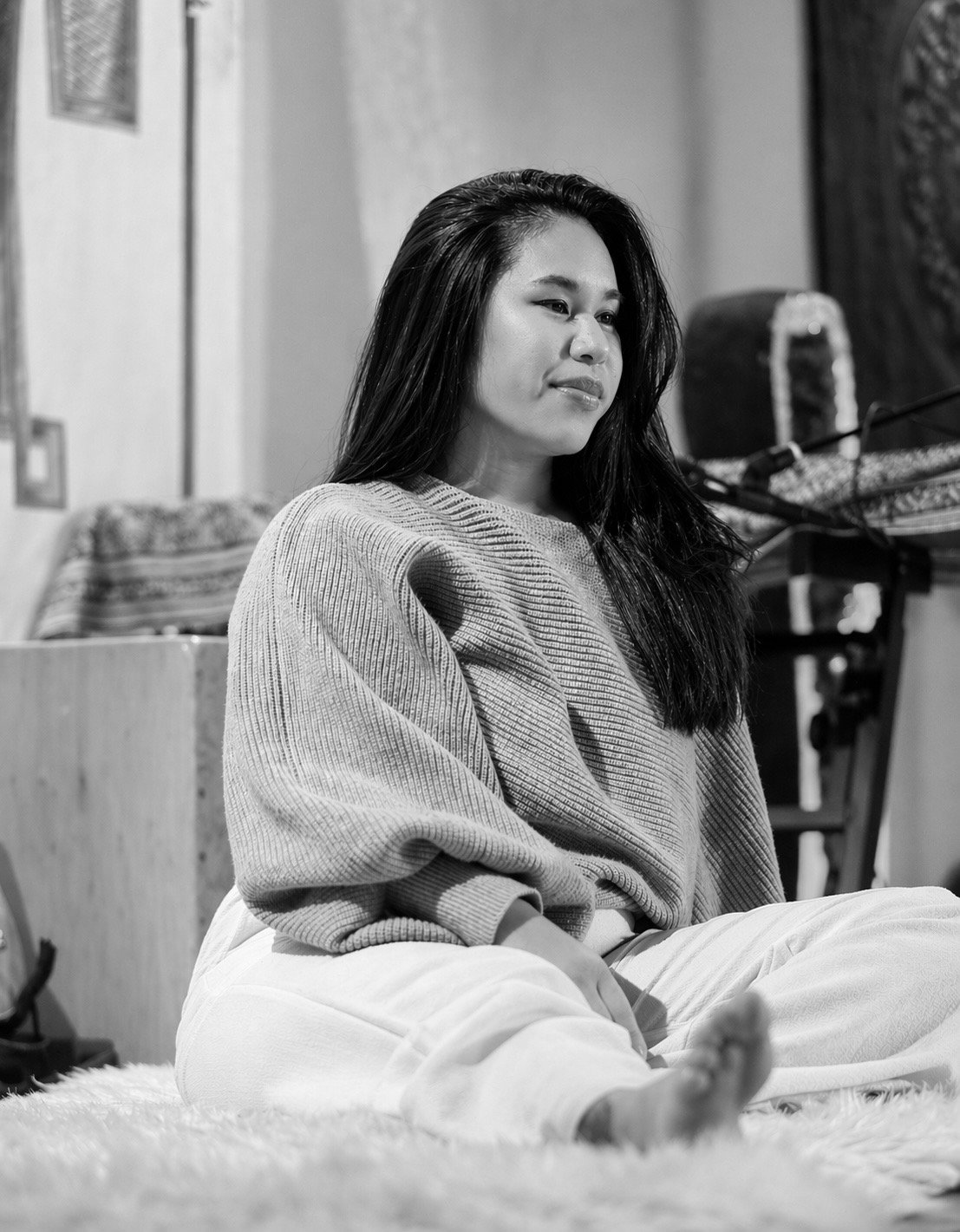 A young woman with long dark hair sitting cross-legged on a soft surface, gazing thoughtfully in a room with patterned decorations and musical instruments in the background, in black and white.