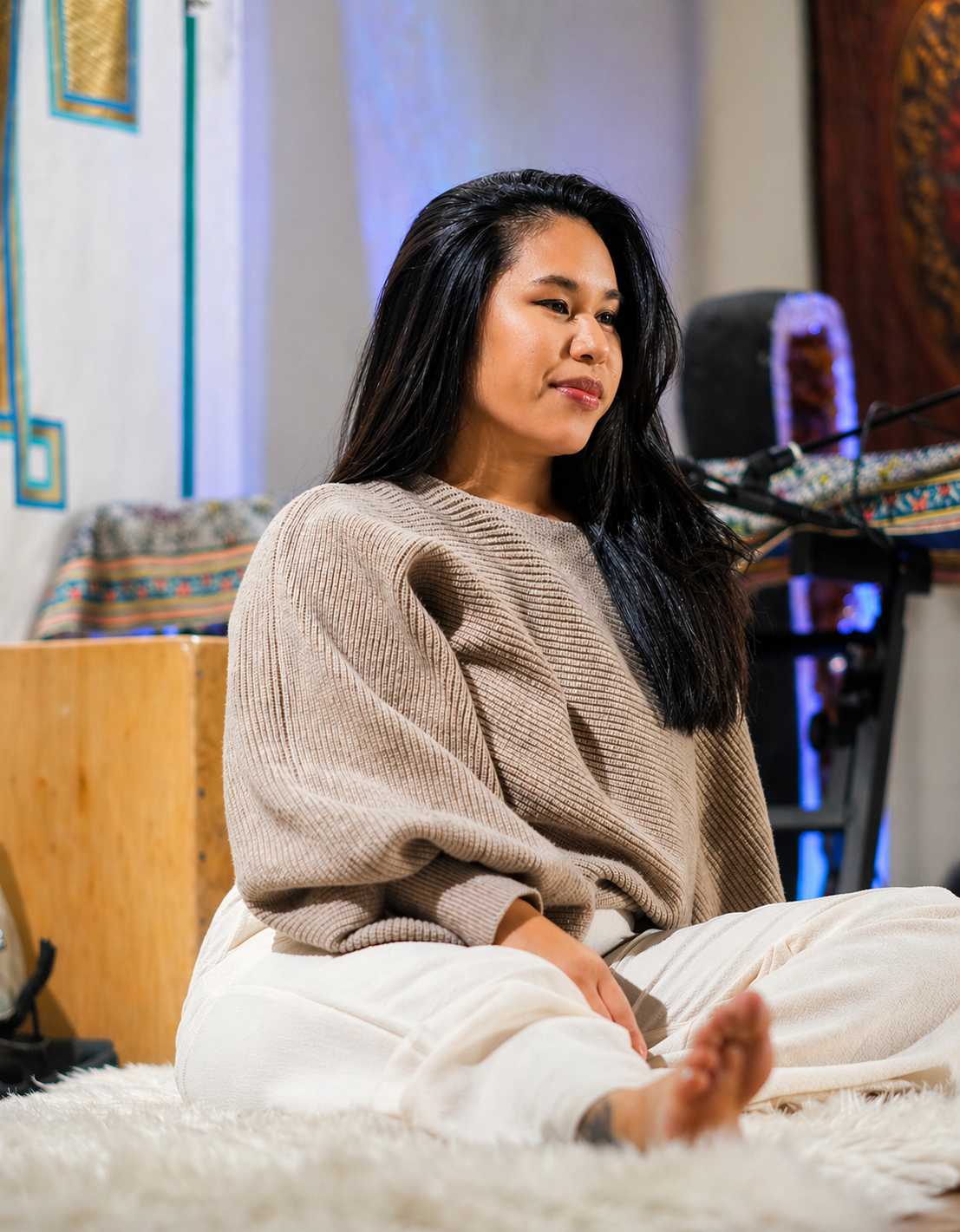 A woman sitting cross-legged on a fluffy white rug in a room with colorful textile decorations and music equipment in the background.