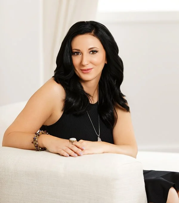 A woman with long black hair, wearing a black sleeveless top and silver jewelry, sitting on a white sofa in a bright room.