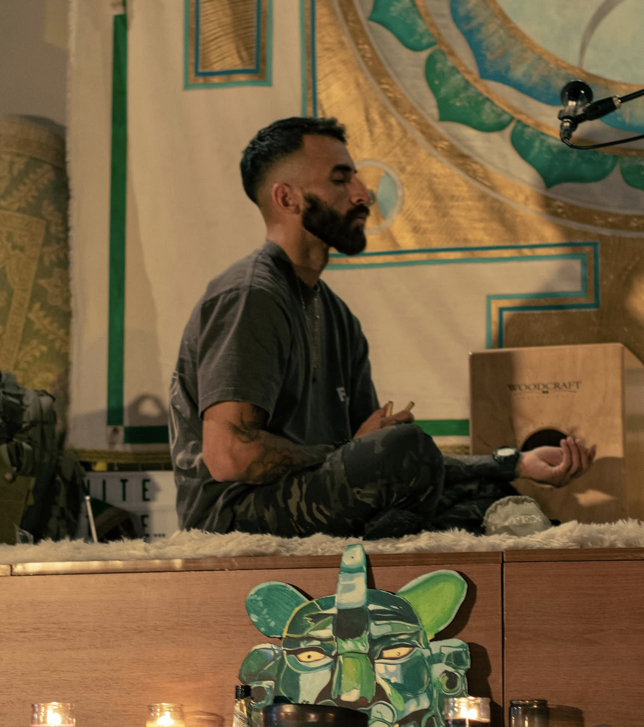 A man with a beard and tattoos on his arm is sitting cross-legged on a furry white rug, holding a small object in his hand and eyes closed, appearing to meditate. He is in front of a wooden box with the word 'Woodcraft' on it and a colorful carved animal head decoration on a wooden surface. The background features a large, colorful mural with abstract patterns.