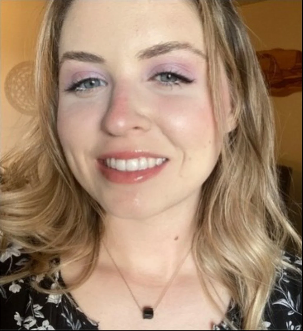 Close-up of a woman with blonde, wavy hair, wearing makeup, smiling, and wearing a black floral dress with a black pendant necklace.