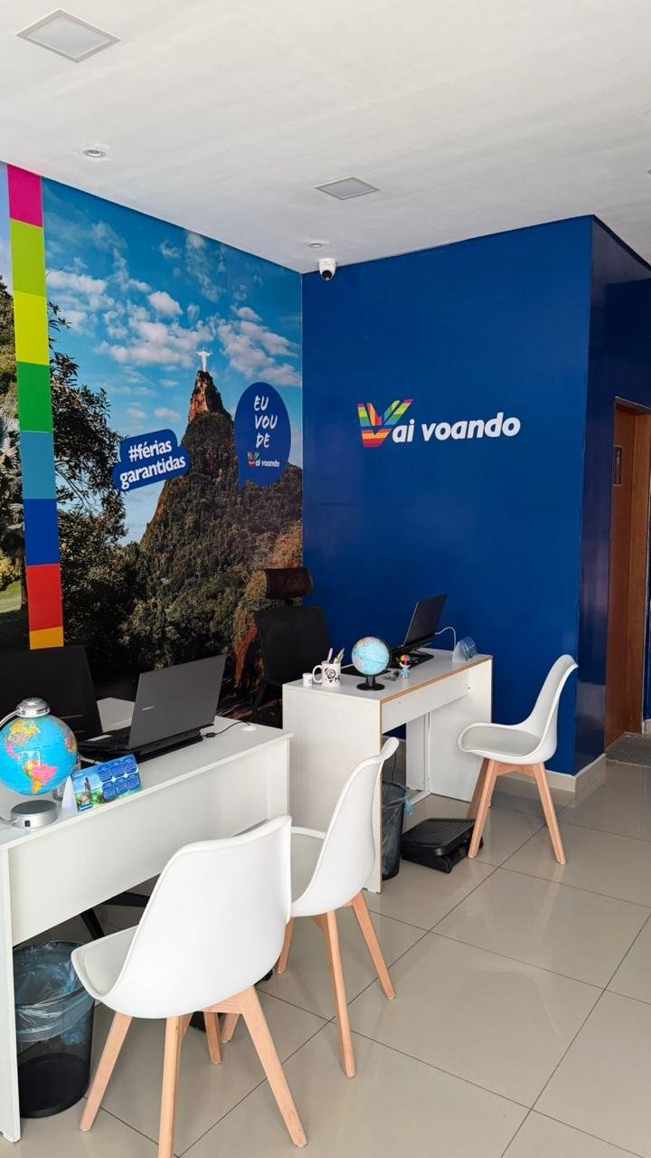 Travel agency office with desks, laptops, and globes, wall mural of a mountain with a cross at the peak, and branding for 'ai voando'