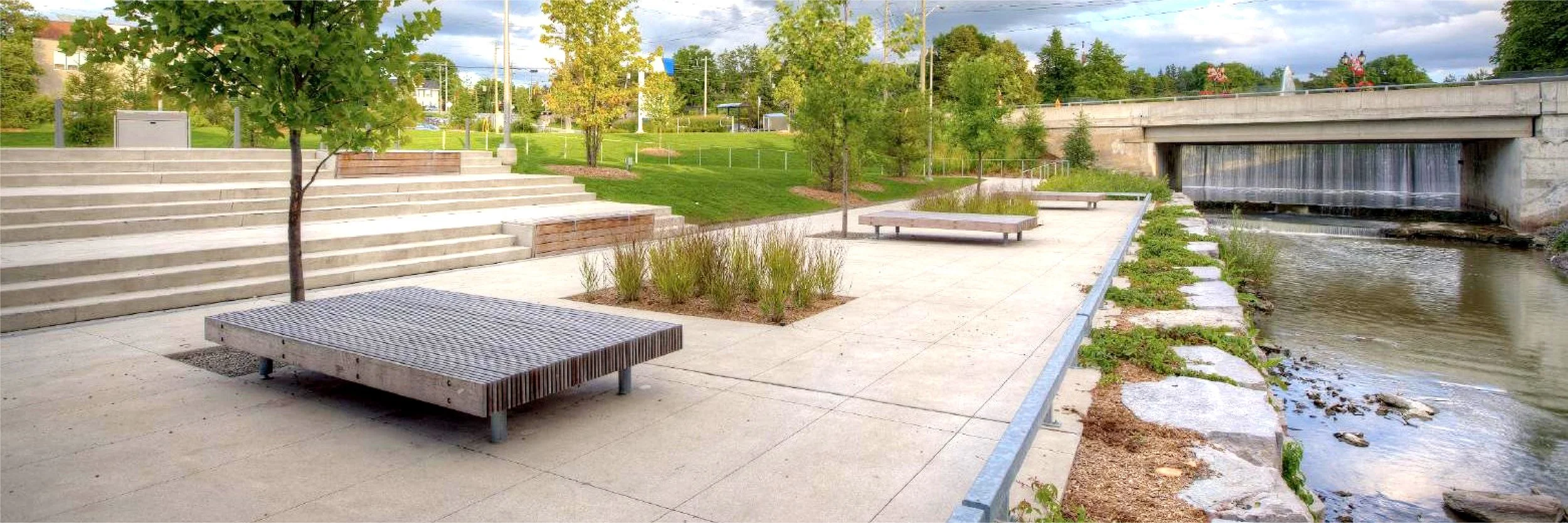 A view of Newarket's Riverwark Commons with concrete stairs, benches, trees, and a water feature.