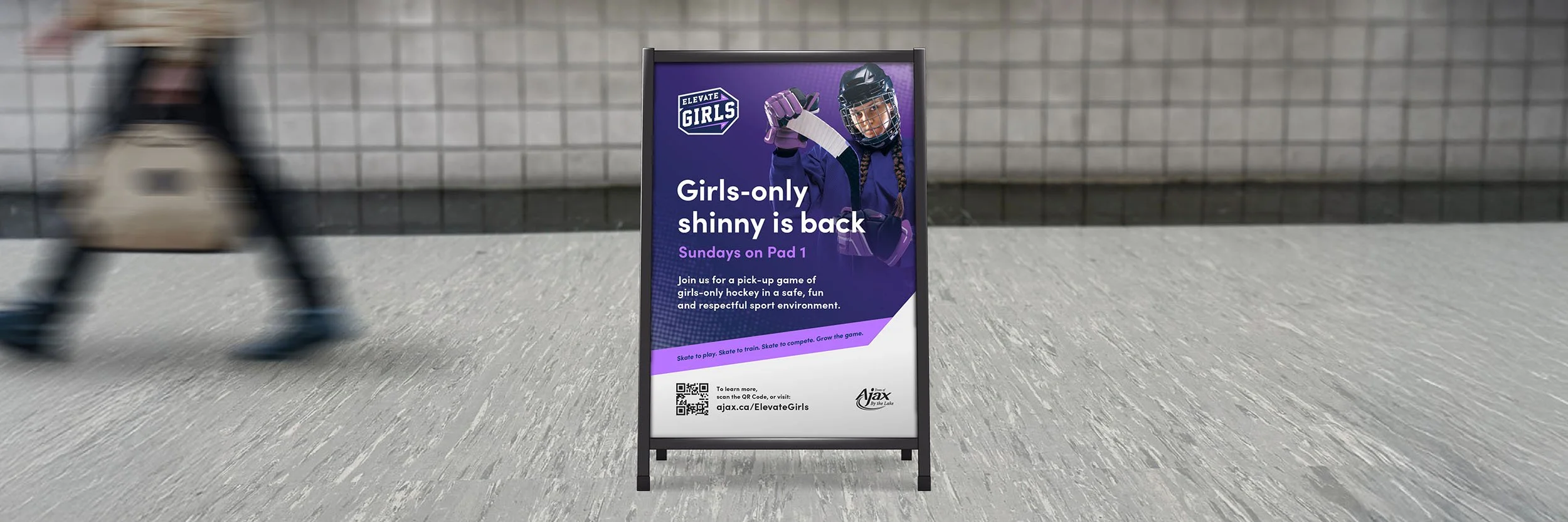 An outdoor sidewalk with a blurred person walking past a black frame signboard advertising girls-only hockey sessions called 'Elevate Girls'. The sign features an image of a girl in hockey gear, holding a hockey stick, and the text states that girls-